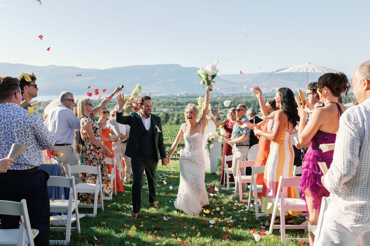 Newlyweds walk back down aisle as guests toss colorful flower petals at outdoor vineyard wedding.