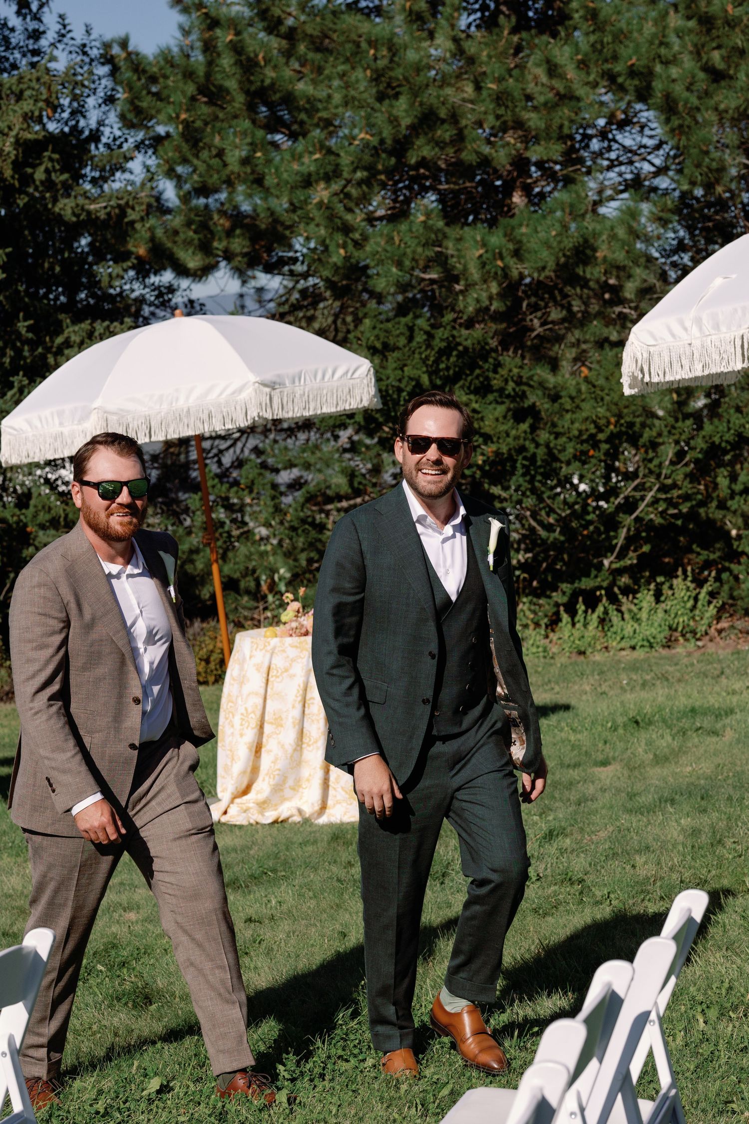 Two groomsmen in dark suits and sunglasses smiling as they walk down outdoor wedding ceremony aisle.