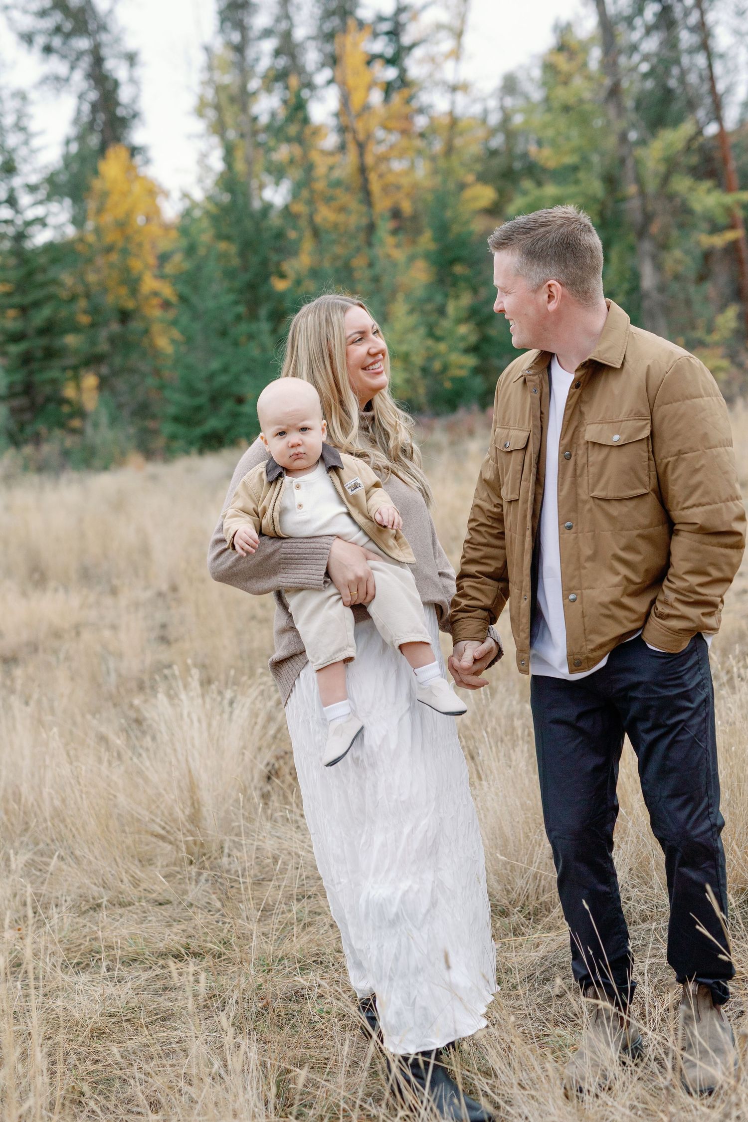 A young family with a baby enjoys an autumn outdoor stroll through a golden field surrounded by evergreen trees.