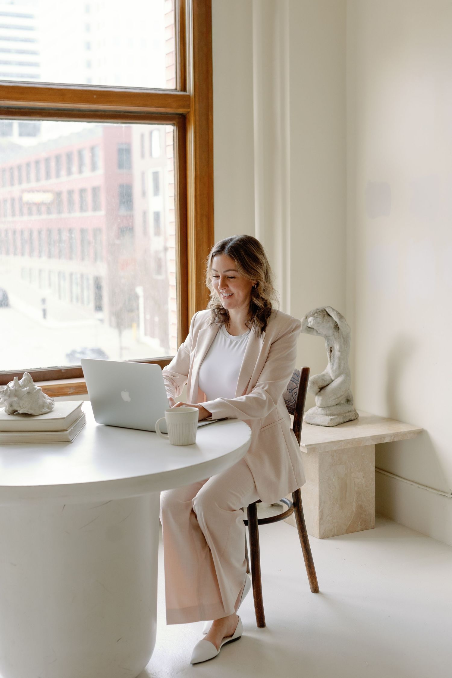 Woman in beige suit smiling at laptop on white round desk by large window in bright modern office.