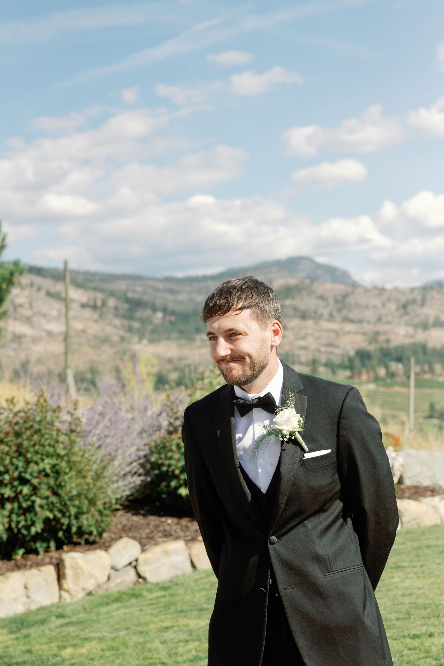 Groom in black tuxedo with bow tie smiles outdoors with scenic mountain landscape behind him.