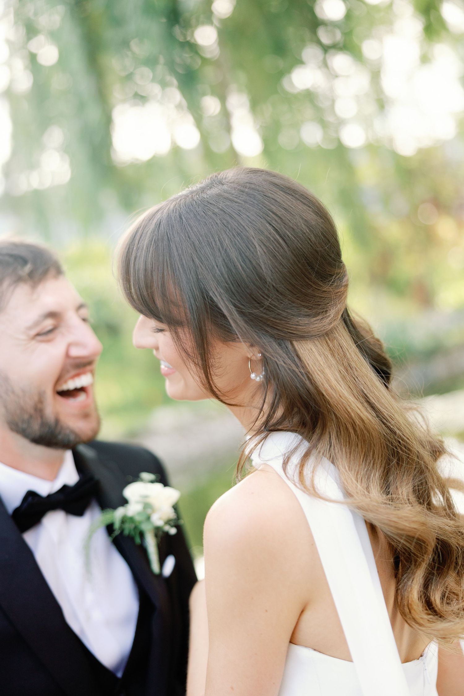 Laughing bride and groom share a joyful moment together outdoors surrounded by soft green bokeh background.