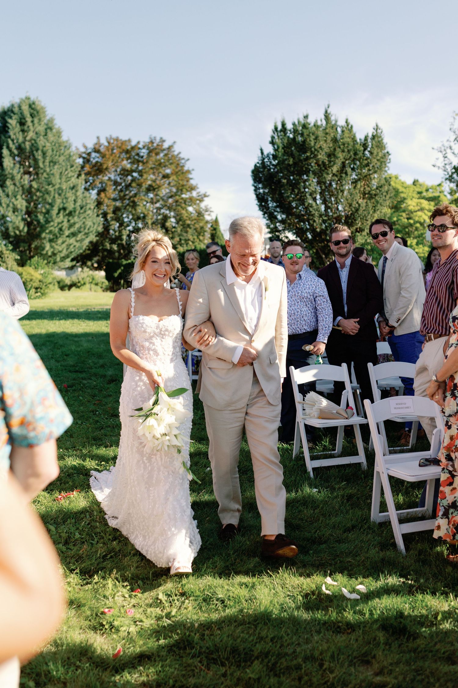 Bride in lace gown walking down the aisle with her father at a sunny outdoor wedding.