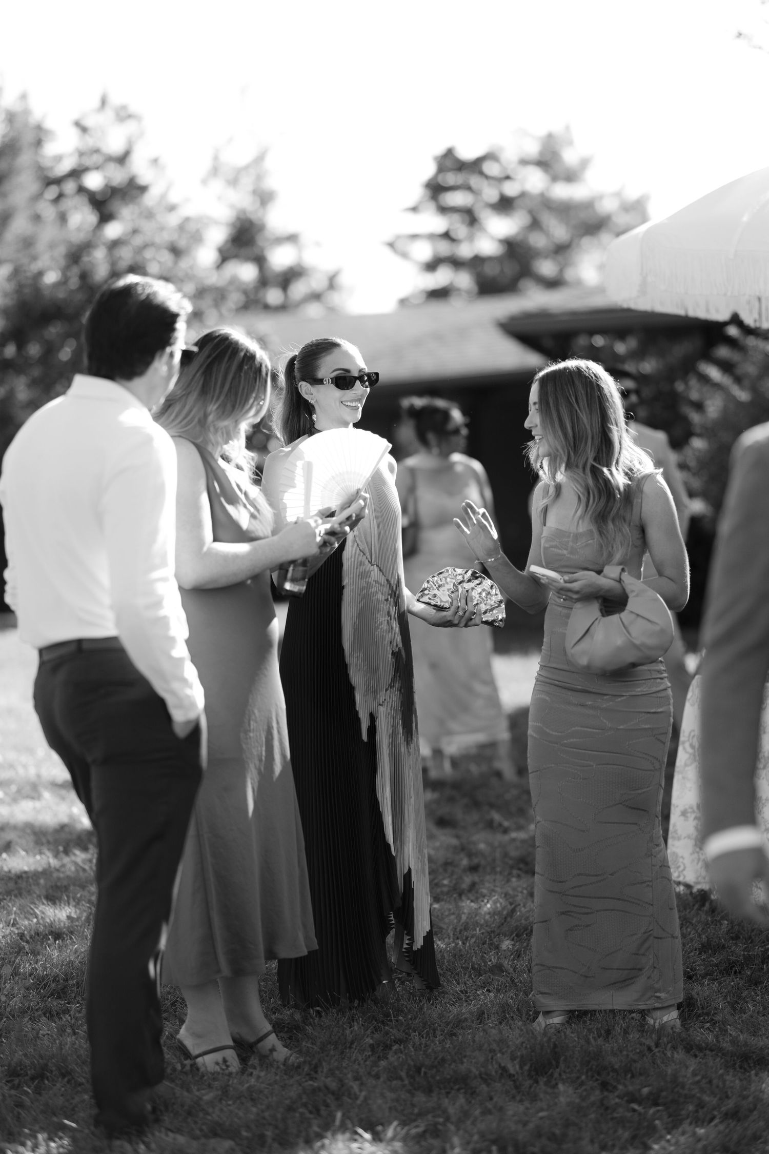 Black and white photo of wedding guests socializing outdoors in elegant attire on sunny day.