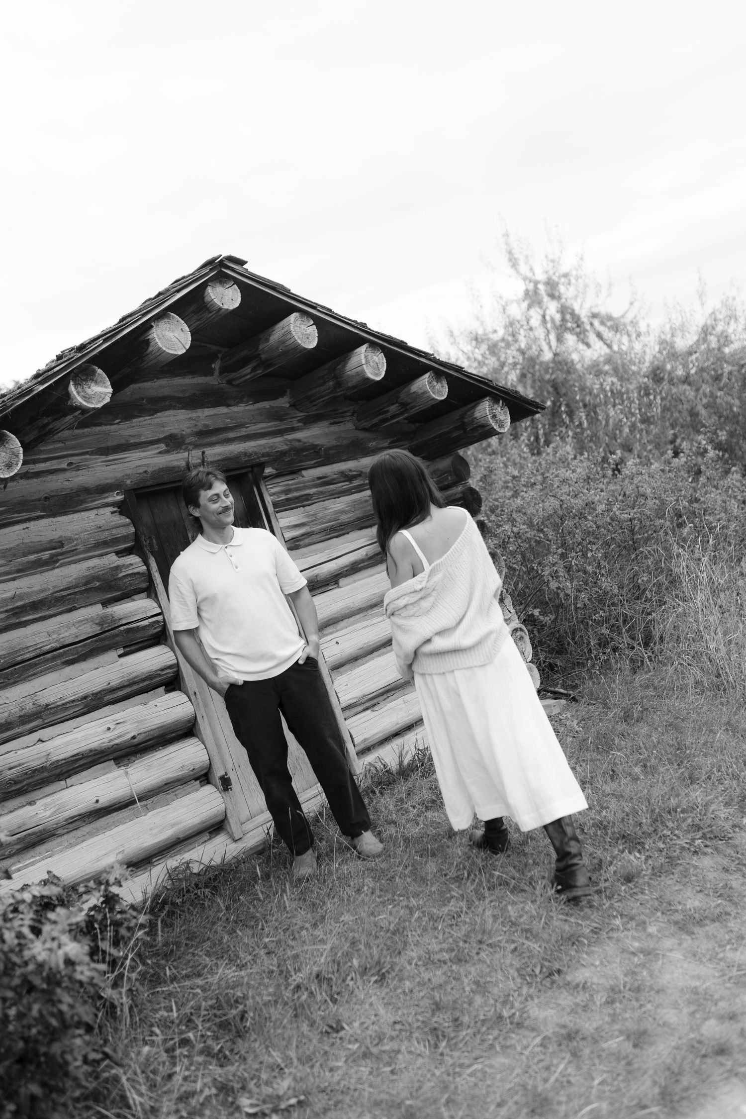 Black and white photo of couple conversing outside a rustic log cabin surrounded by wild vegetation.