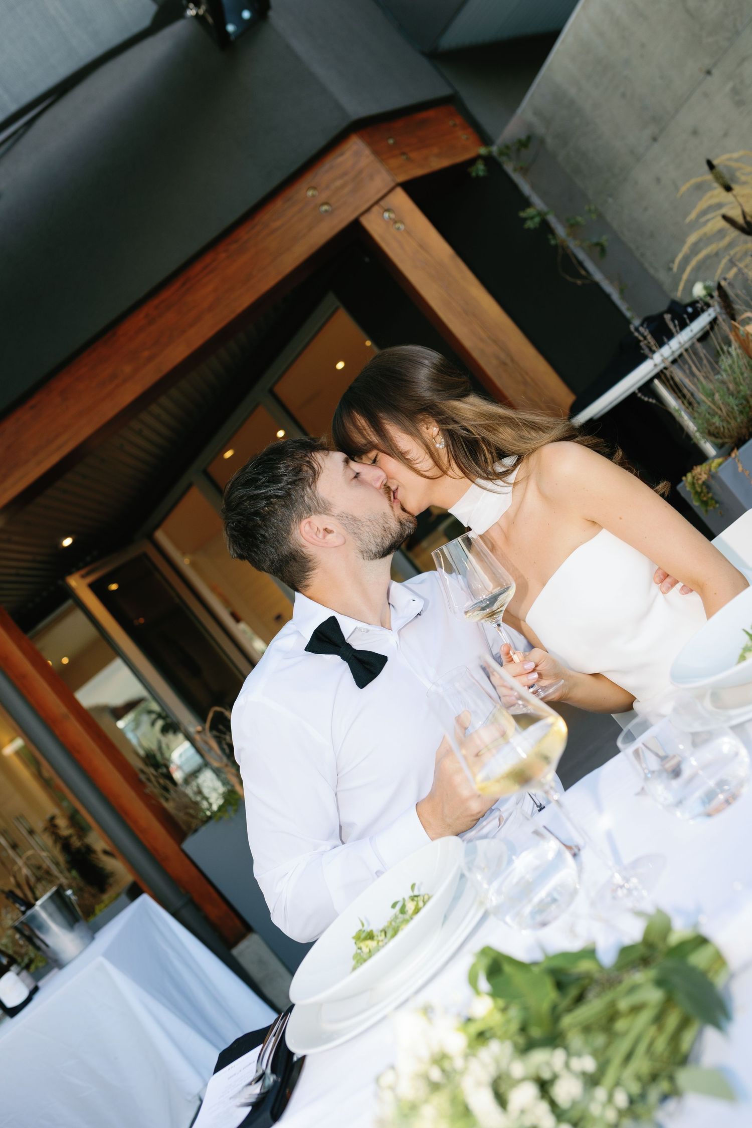 Bride and groom sharing a kiss at wedding reception table with wine glasses nearby.