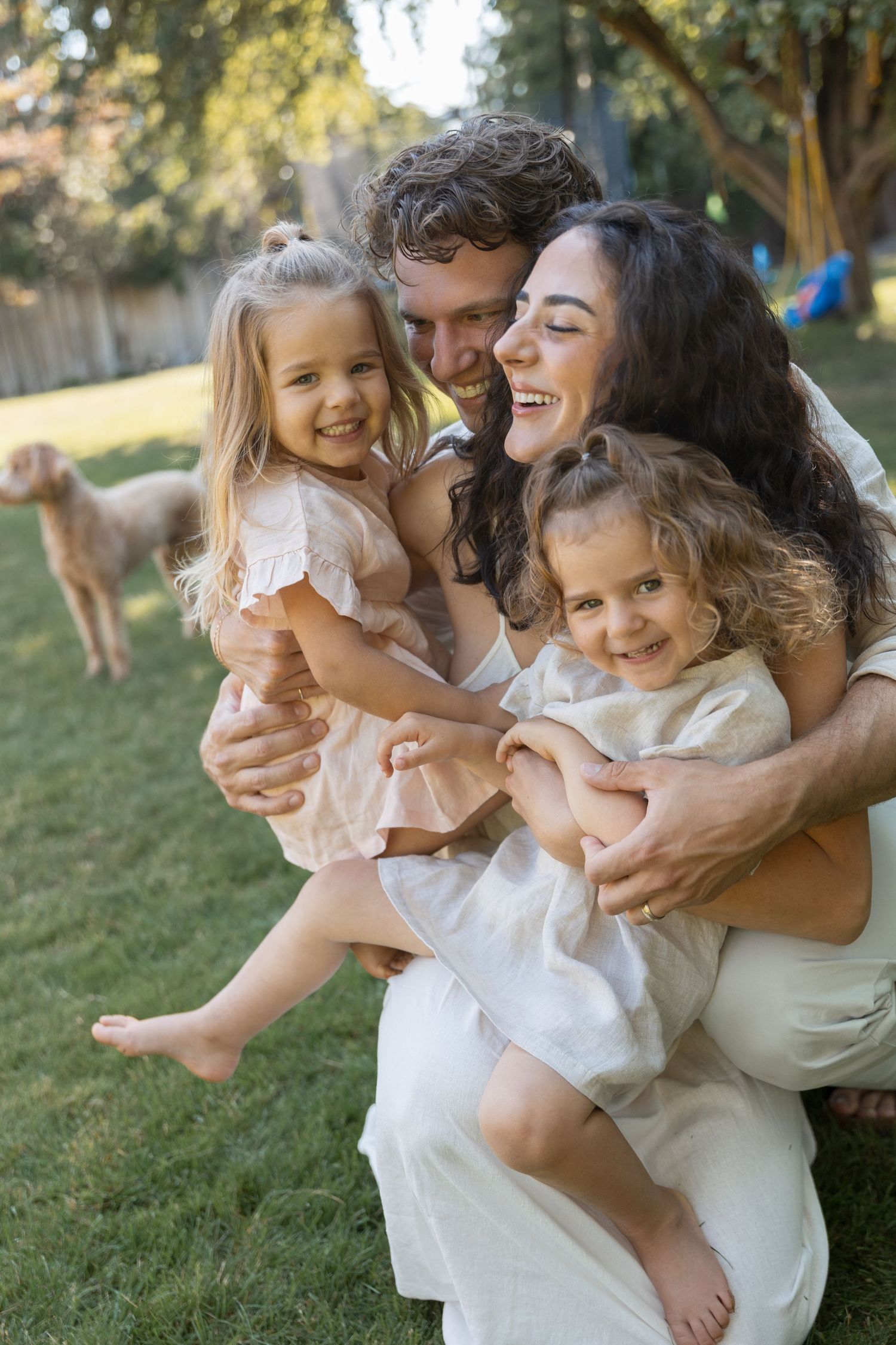 Happy family with two young daughters laughing and embracing outdoors in a sunny backyard with a dog in background.