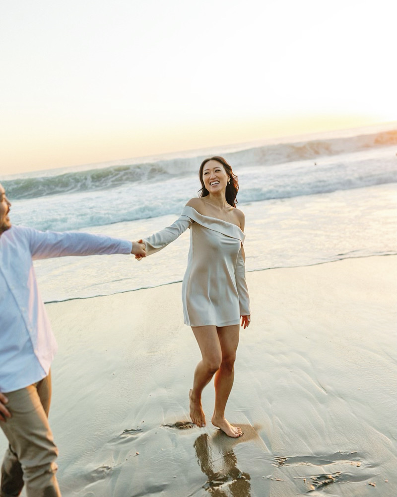 A smiling woman in an off-shoulder dress holds a mans hand while walking along a sunny beach at sunset.