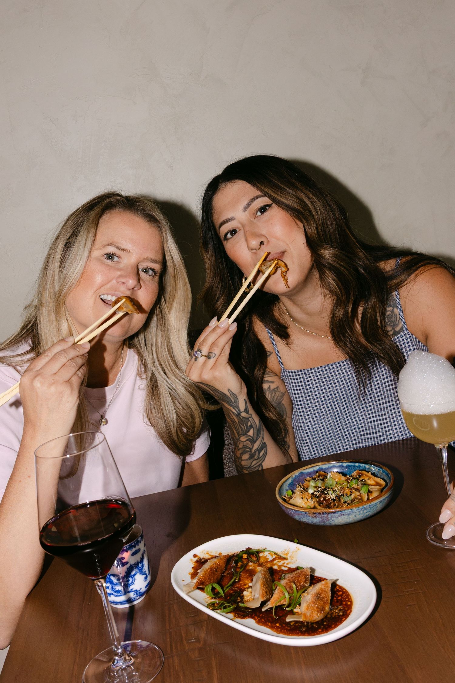 Two women smile while eating with chopsticks at a restaurant table with Asian dishes, wine, and beer.