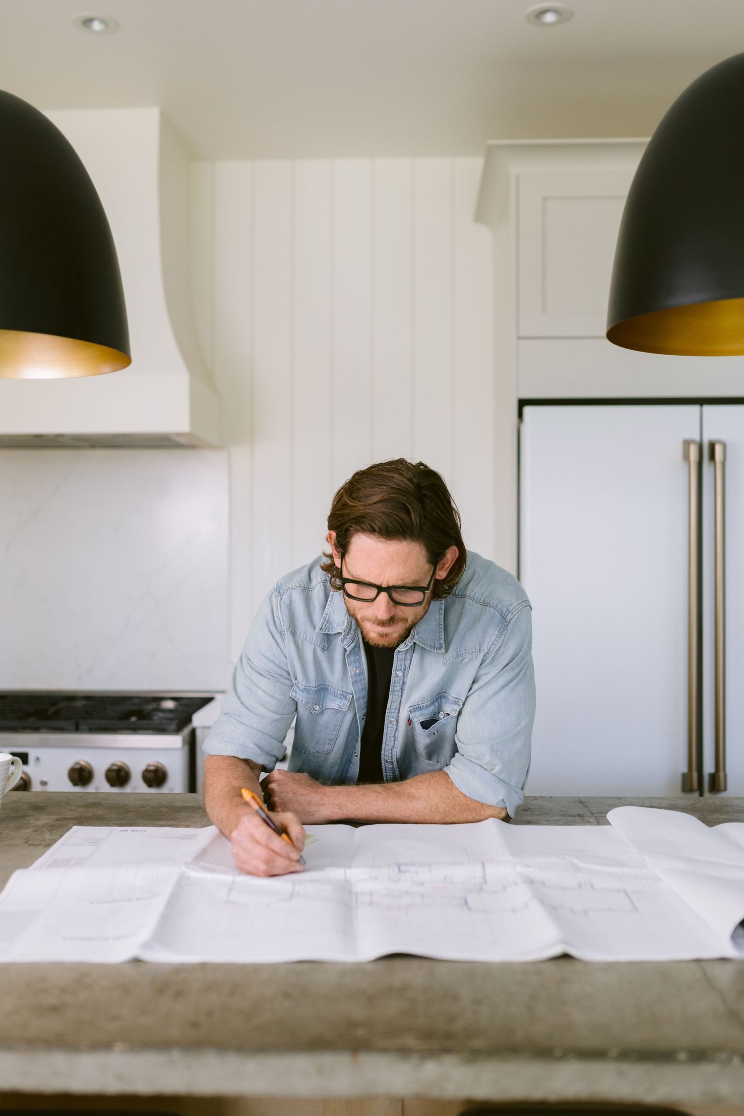 Man in glasses reviewing architectural blueprints at a kitchen counter with modern pendant lights overhead.