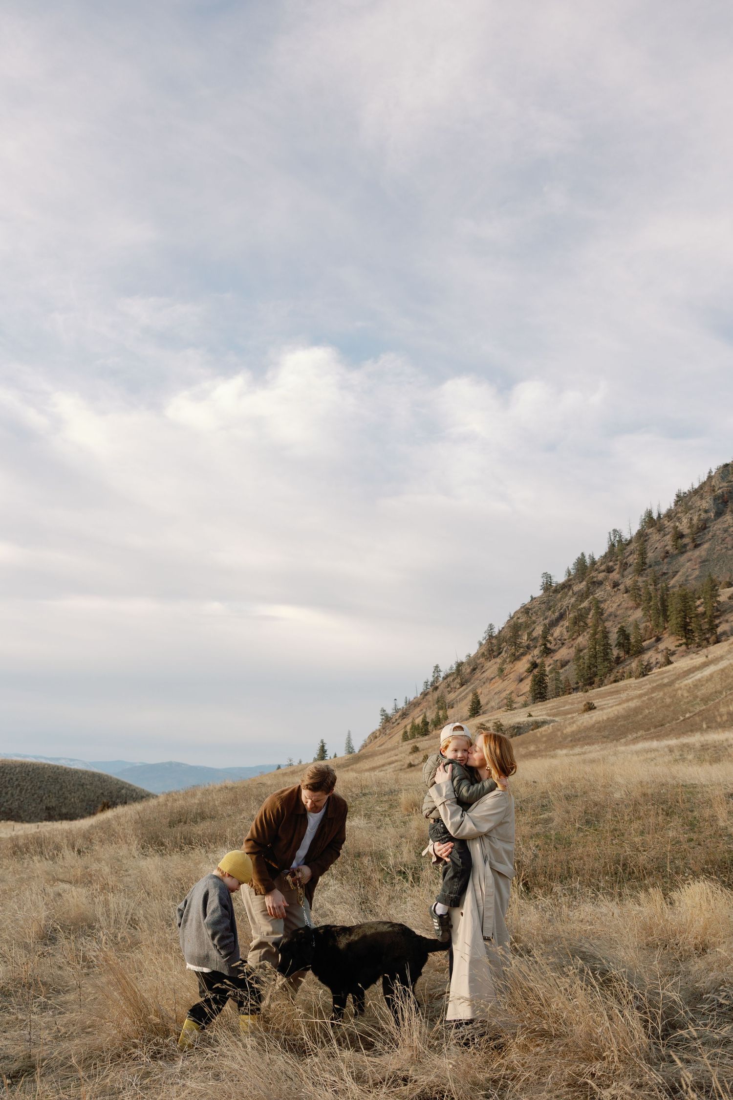 Family with young children and black dog enjoying outdoor hike on golden hillside meadow with mountain backdrop.