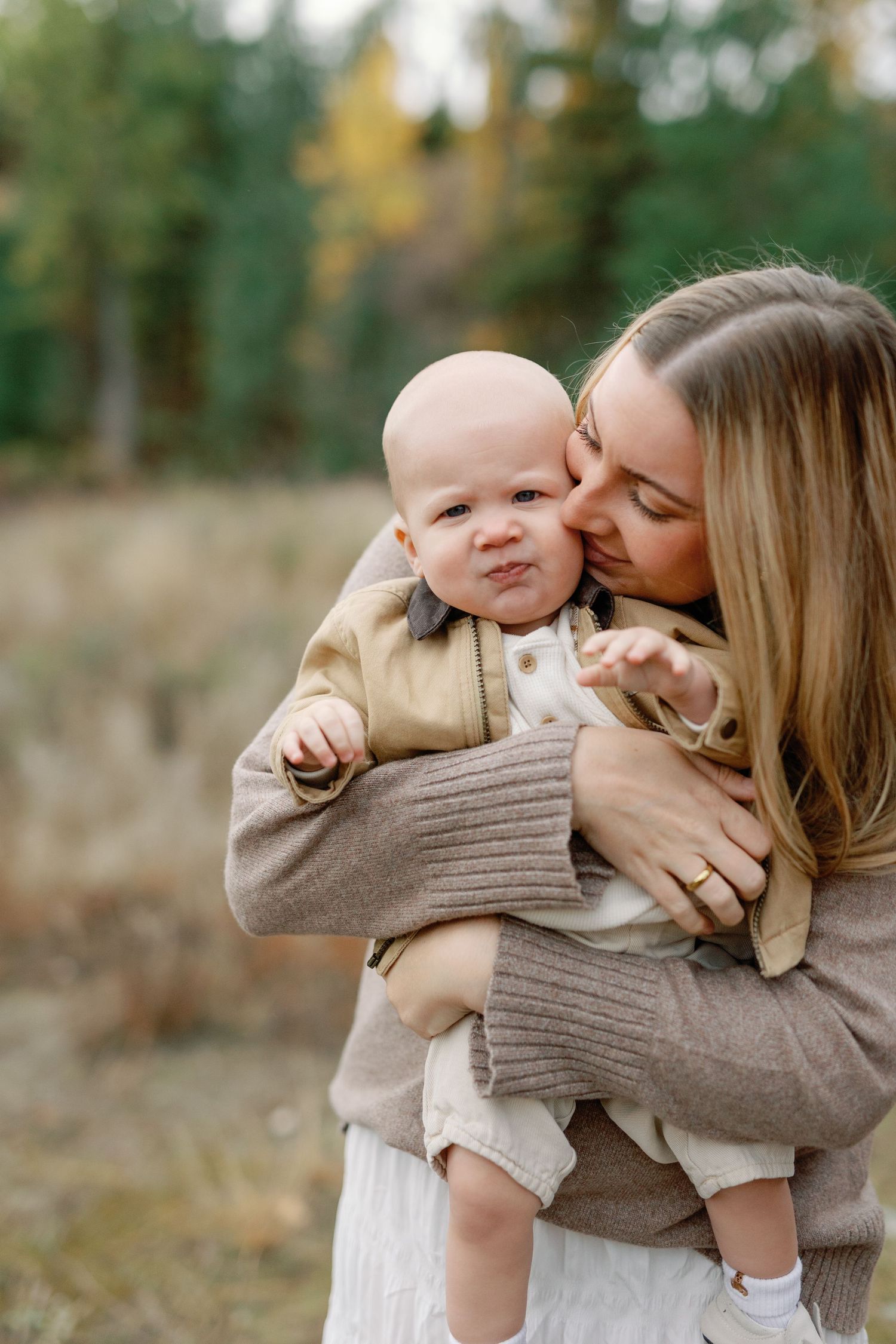 A loving mother kisses her baby's cheek while holding them outdoors in an autumn setting.