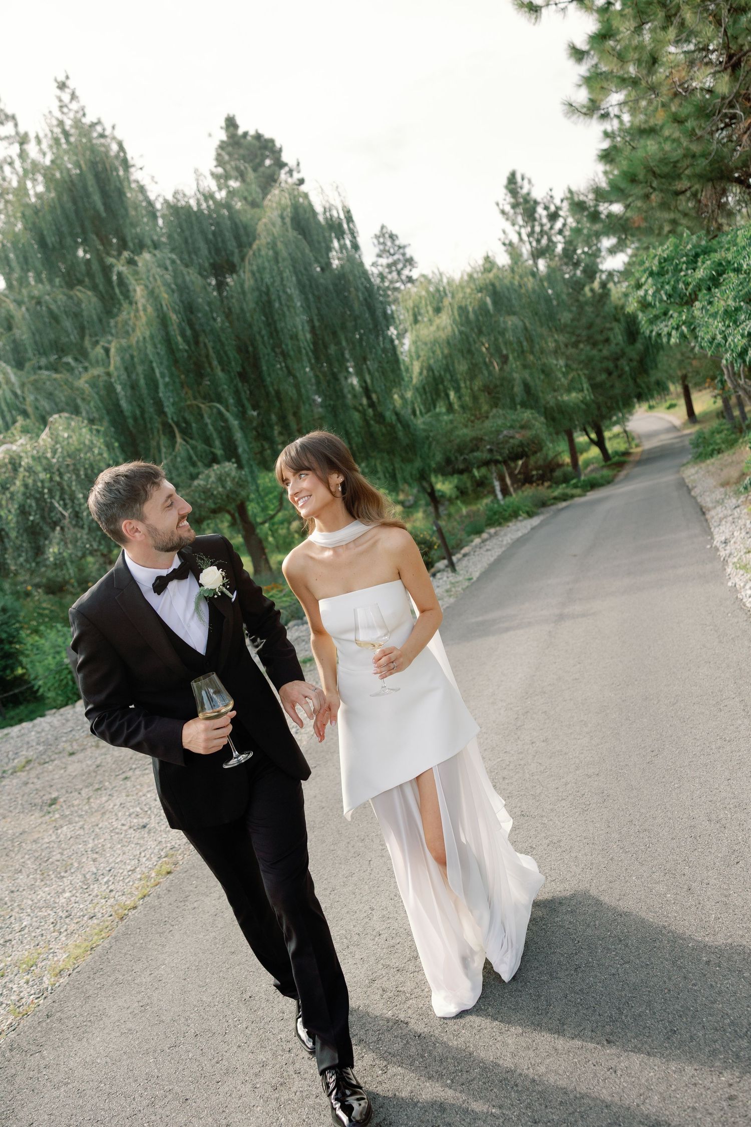 Bride and groom walking hand in hand on a tree-lined path, holding champagne glasses in elegant wedding attire.