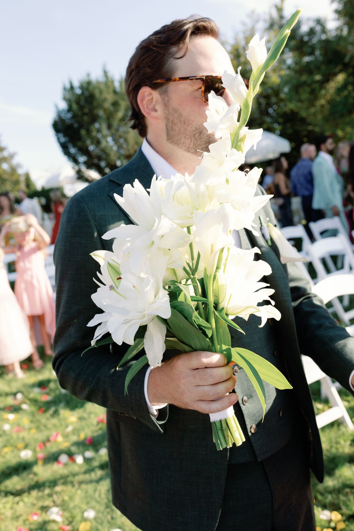 Groomsman in dark suit holds elegant white lily bouquet at outdoor wedding.