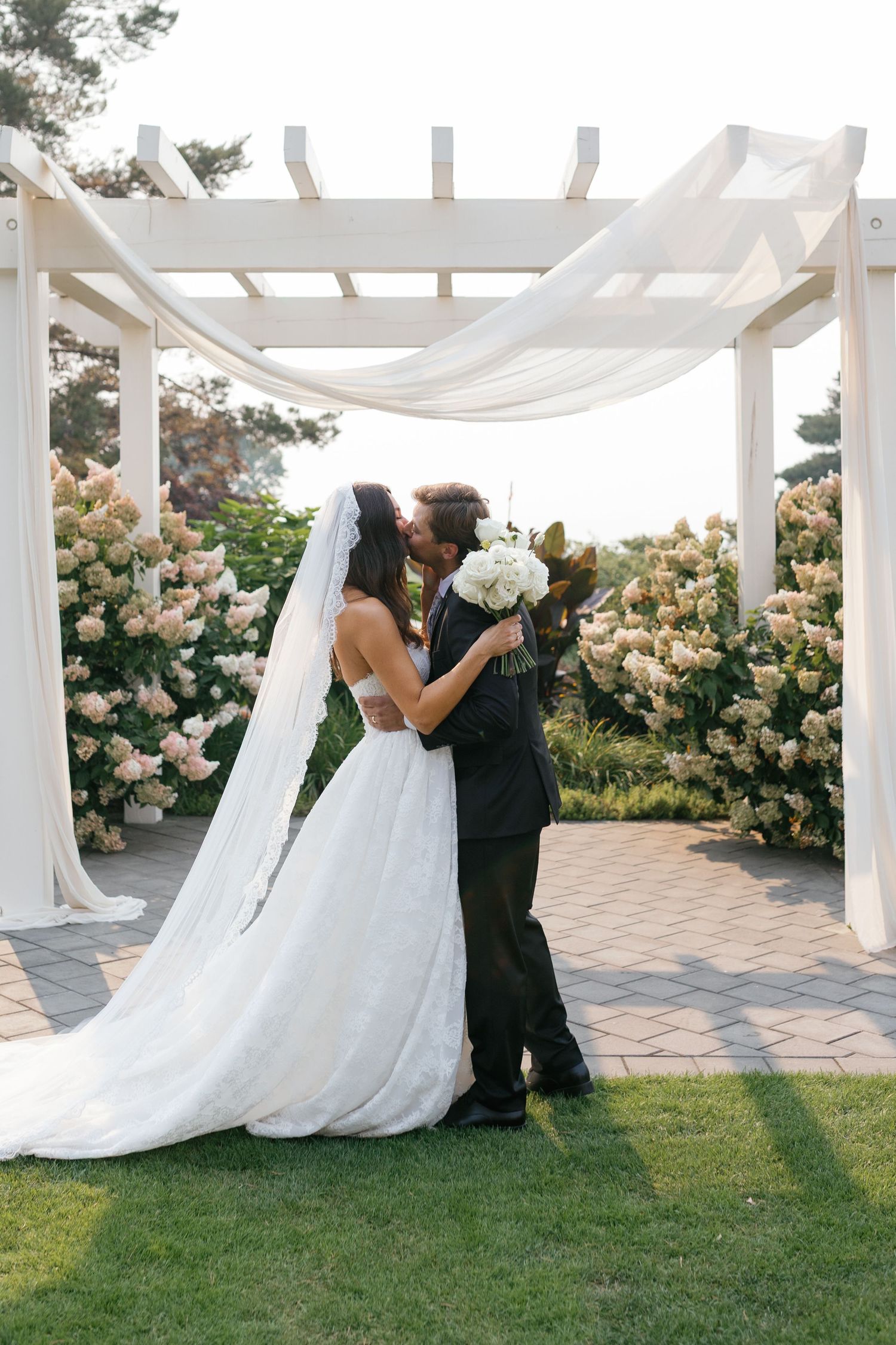 Bride and groom share first kiss under white pergola draped with fabric, surrounded by blooming flowers at outdoor wedding ceremony.
