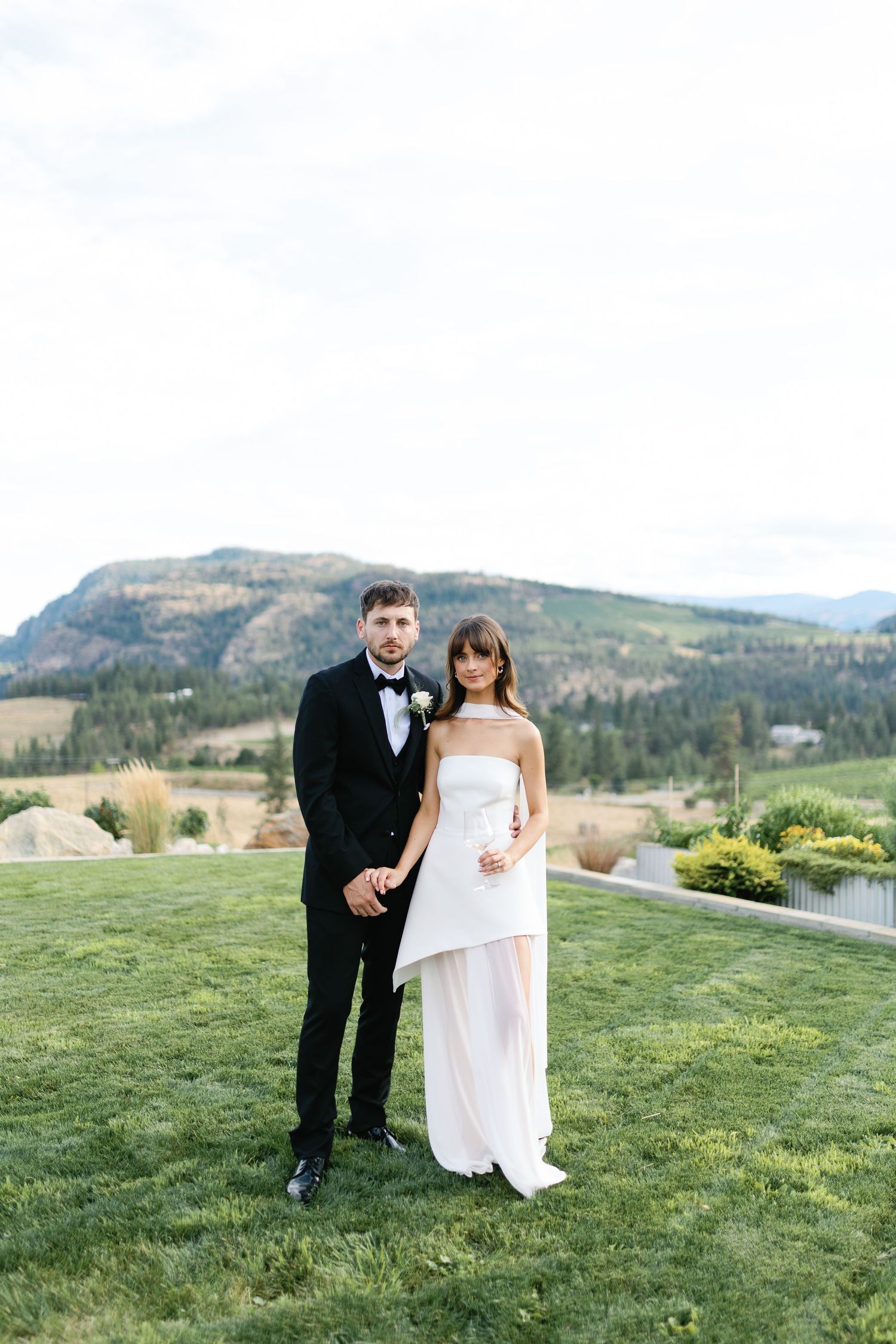 Bride and groom pose on green lawn with mountain backdrop, she in white strapless gown, he in black tuxedo.
