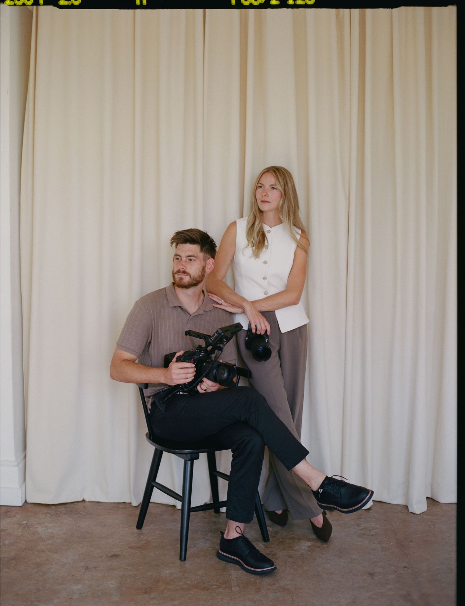 Wedding photography duo posing with professional cameras against cream curtain in film-style portrait.