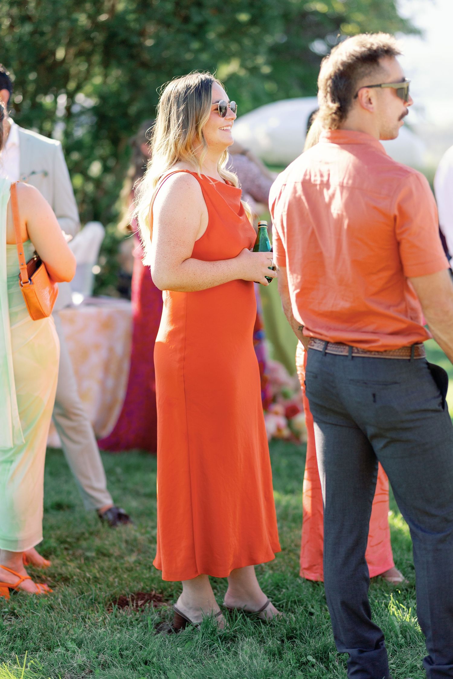 Woman in orange midi dress holding a drink at an outdoor wedding reception on a sunny day.