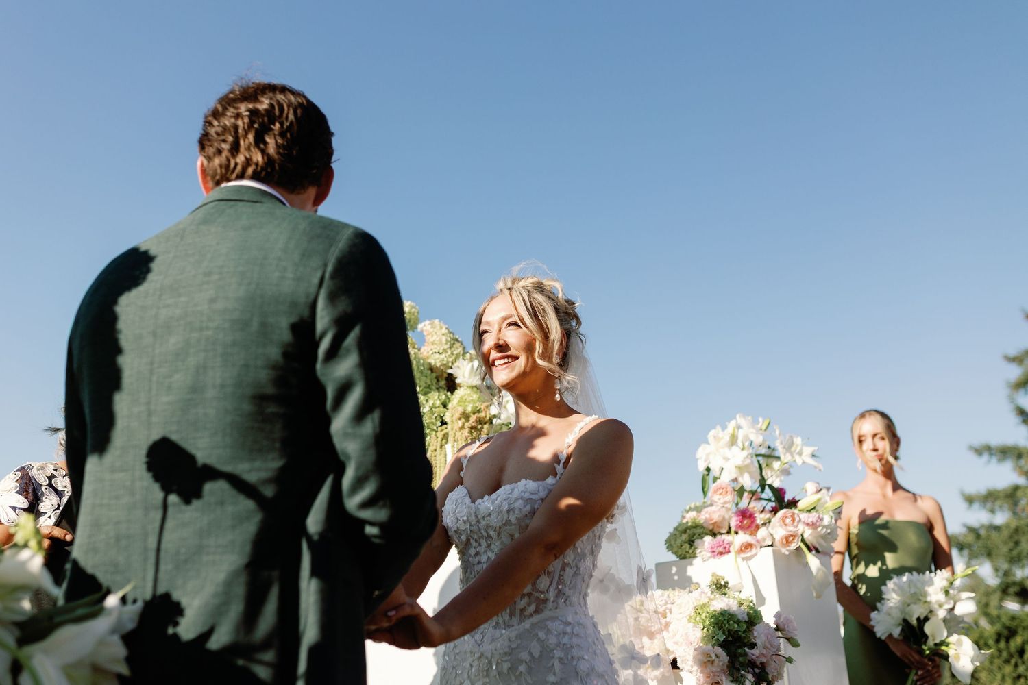 Bride in lace gown smiling radiantly while holding grooms hands during sunny outdoor wedding ceremony.