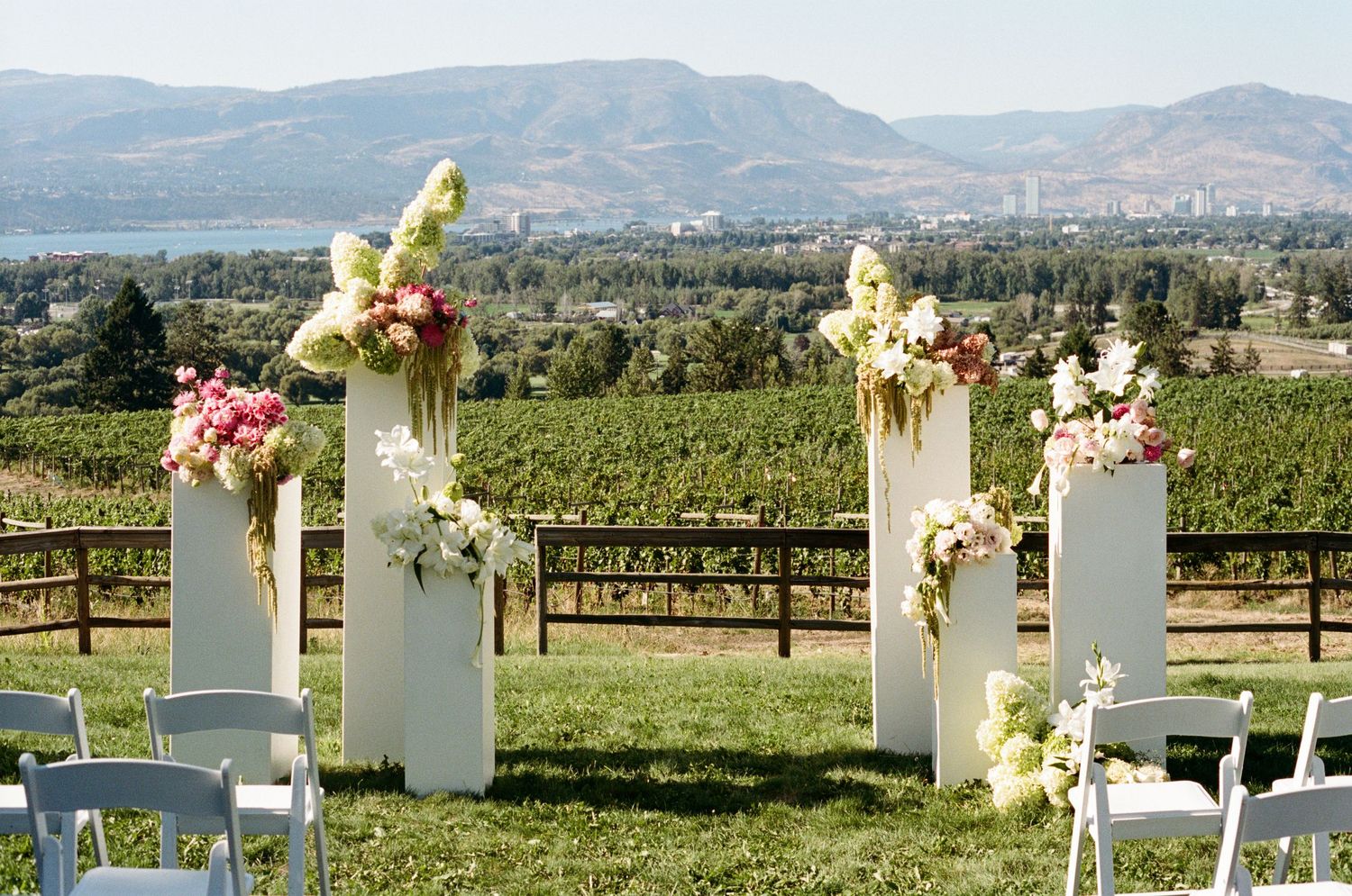 Elegant outdoor wedding ceremony setup with white pillars, floral arrangements, and vineyard views.