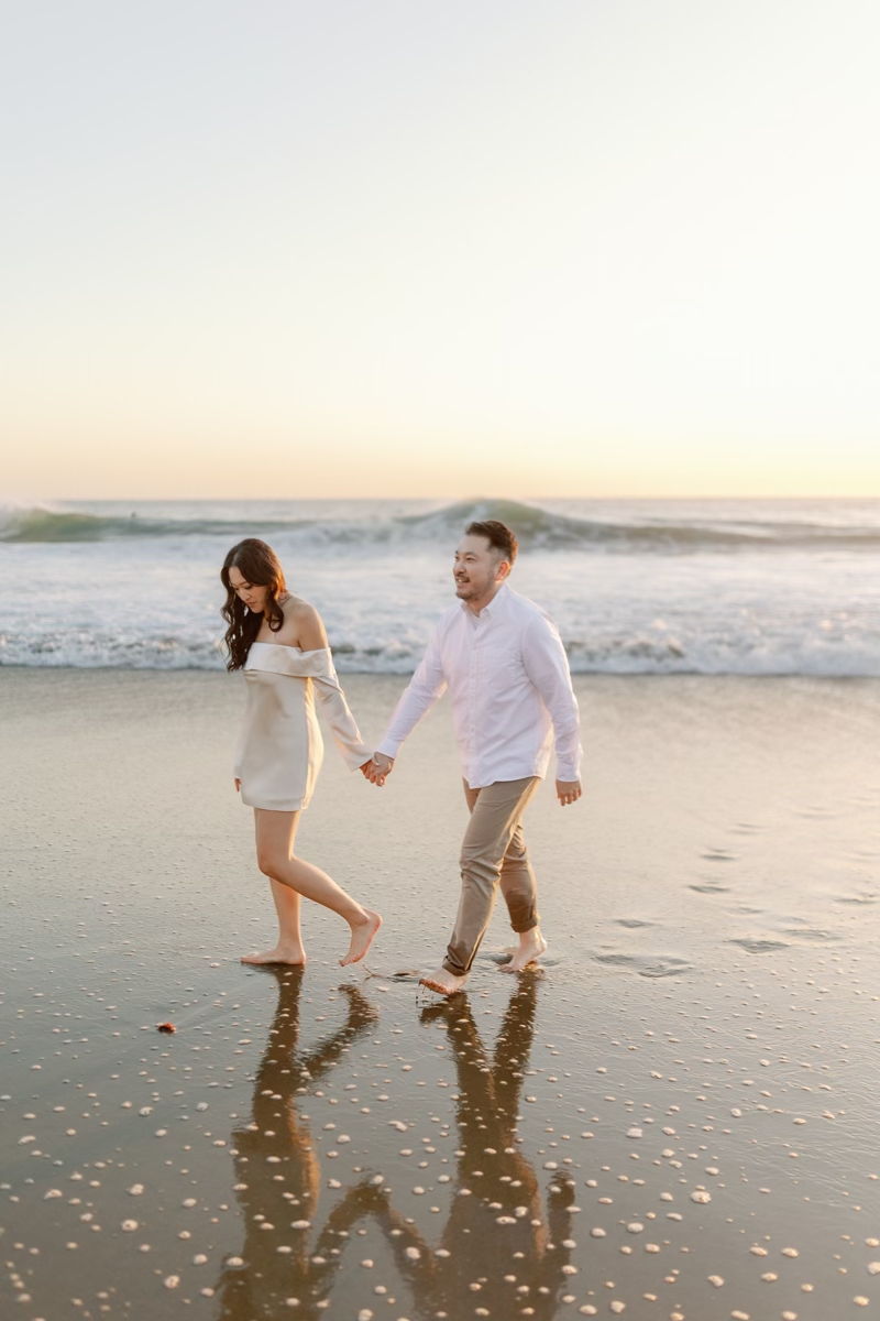 Barefoot couple holding hands and walking along a reflective wet beach at golden hour sunset.