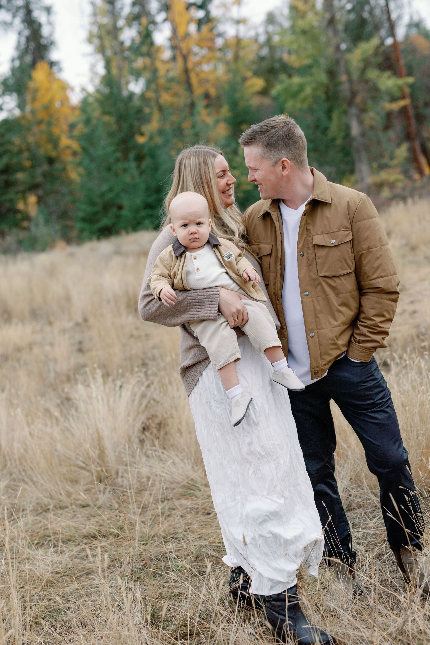 A happy young family with a baby poses in an autumn meadow surrounded by colorful trees in a warm outdoor portrait.