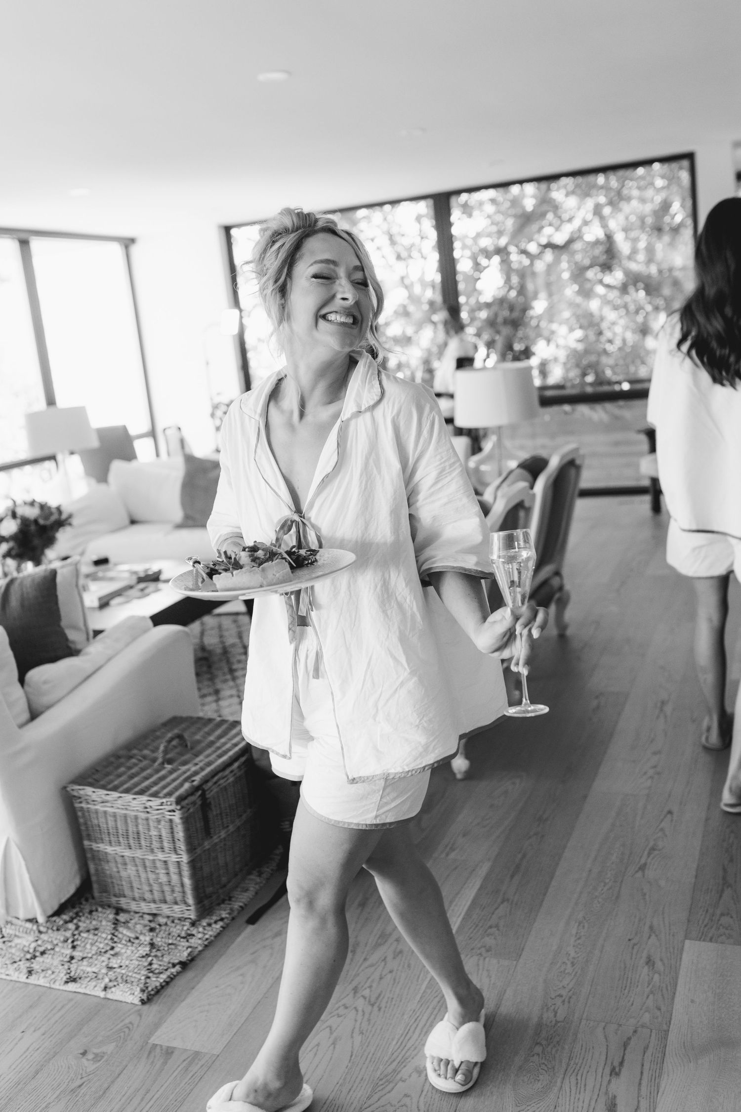 Joyful bride in white pajamas holding champagne and flowers during wedding morning preparations in modern home.