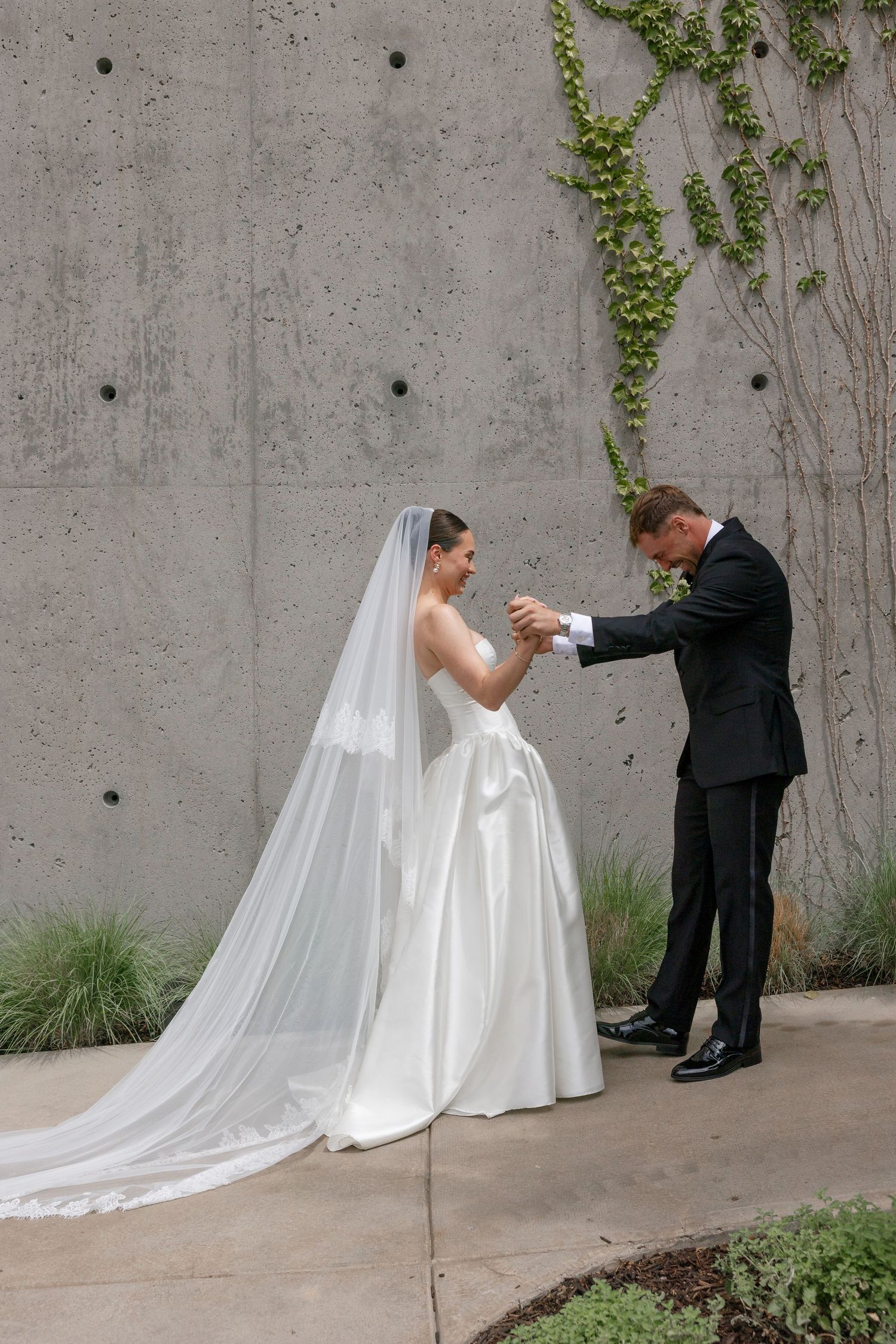 Groom adjusting bride's cuff on wedding day, bride in white gown with long veil, outdoors against concrete wall.