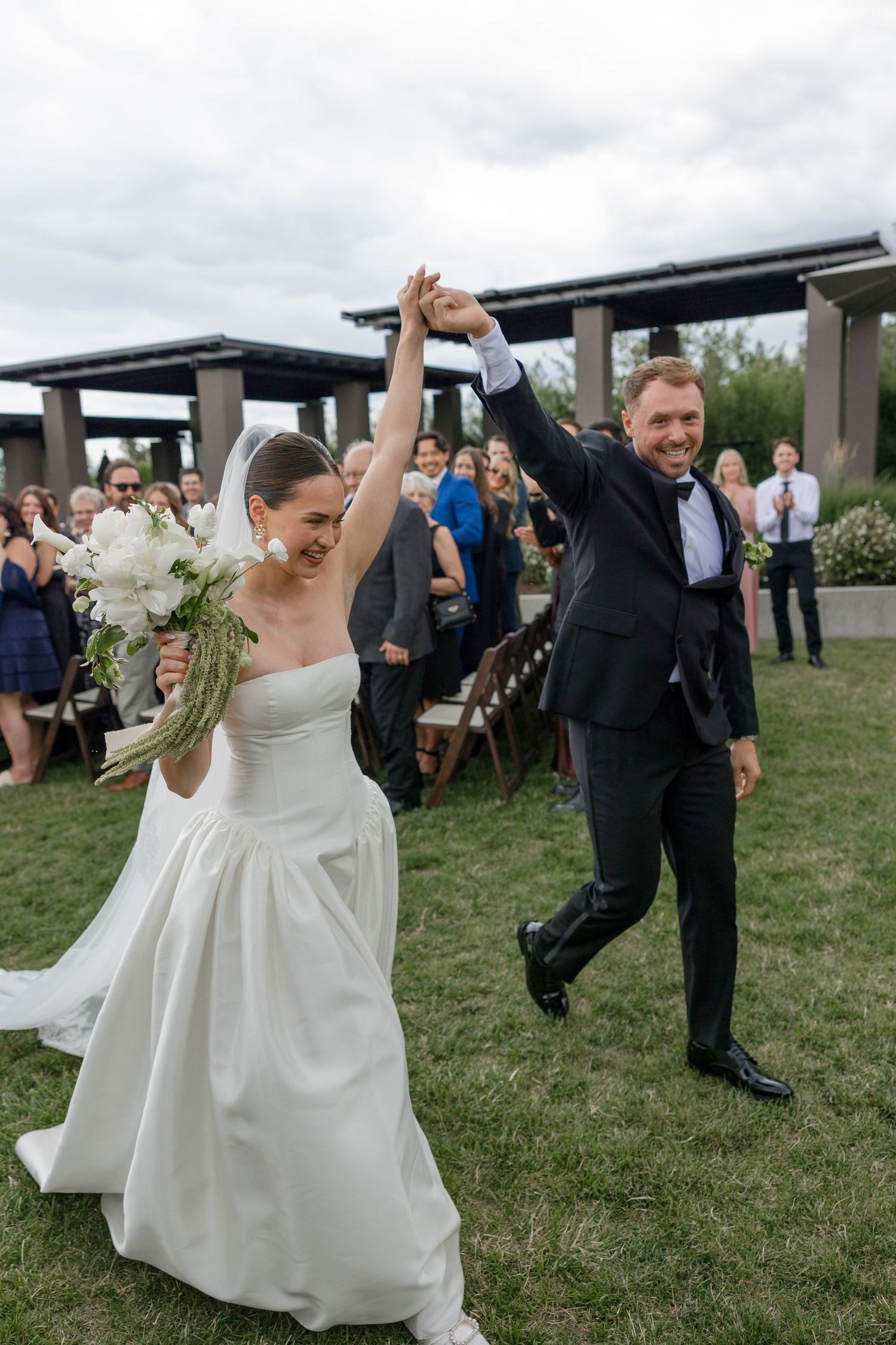 Joyful newlyweds raise hands walking down aisle after outdoor wedding ceremony, bride holding white bouquet in elegant gown.