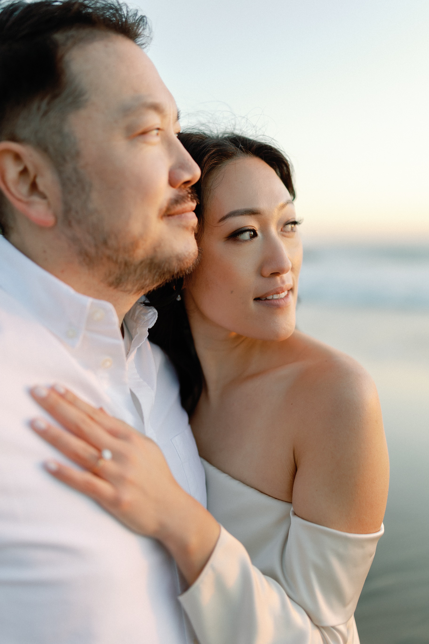 Close-up of engaged couple on beach at sunset, woman showing diamond ring while embracing man in white shirt.
