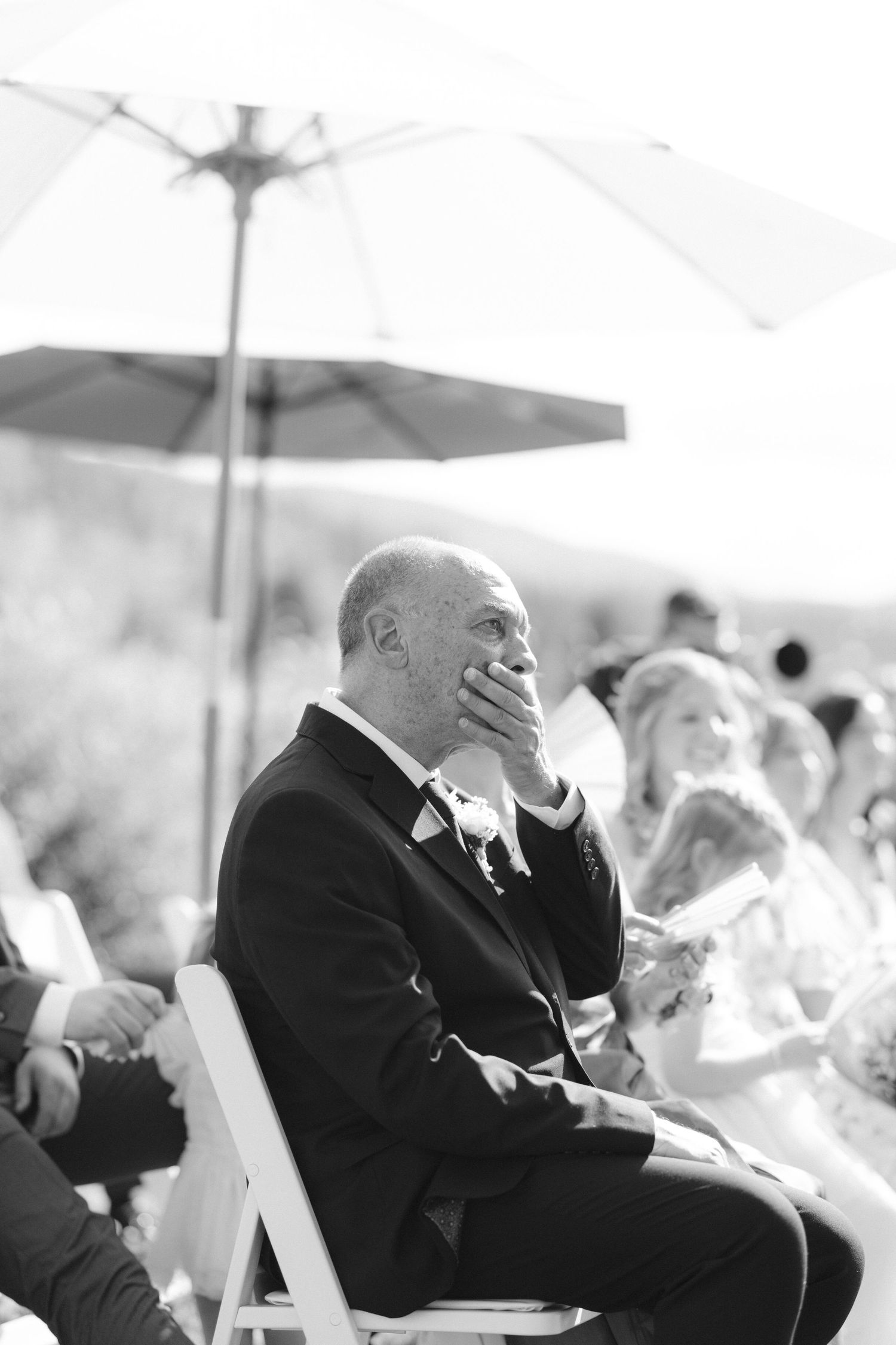 Emotional wedding guest in suit covering mouth with hand during outdoor ceremony.
