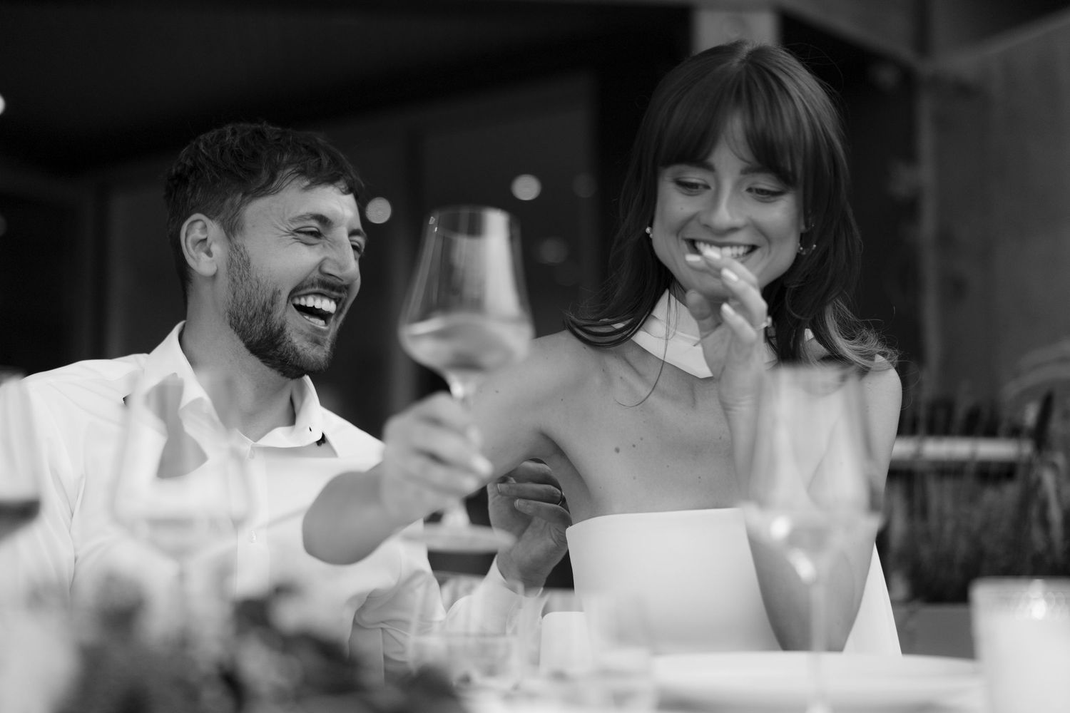 Black and white photo of laughing bride and groom enjoying wine at wedding reception.