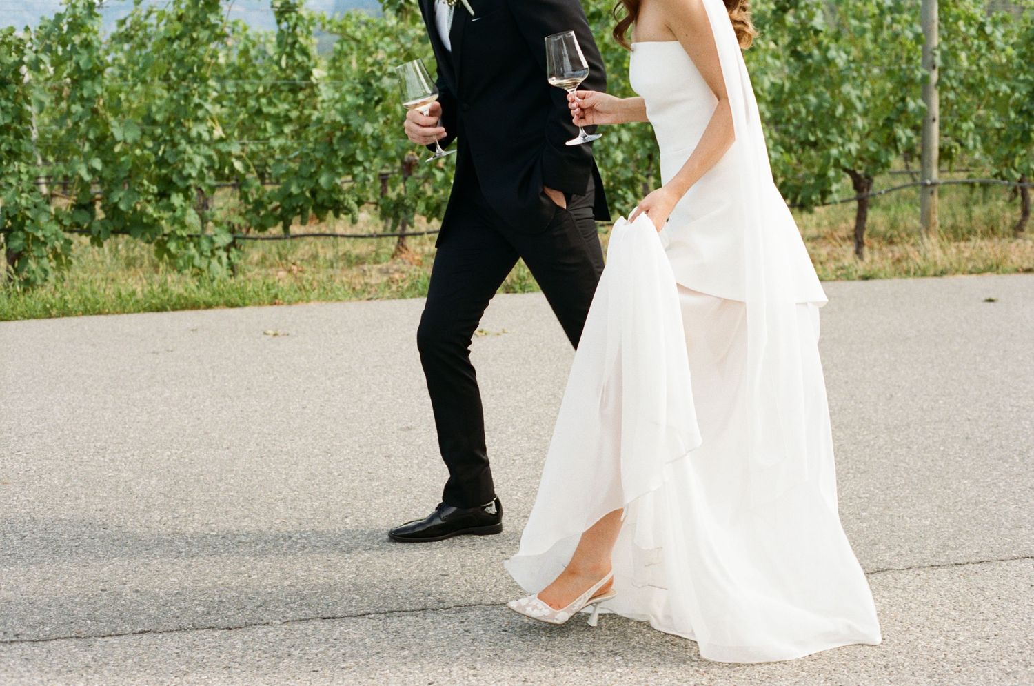 Bride in flowing white gown and groom in black tuxedo walking together at vineyard wedding venue.