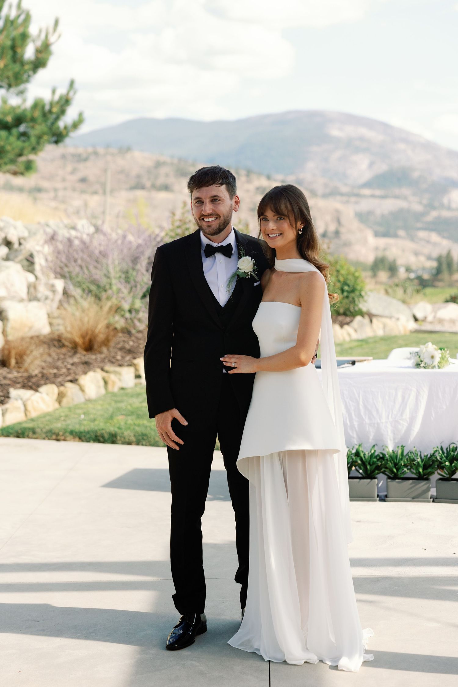 Bride in white halter gown and groom in black tuxedo pose together at outdoor wedding venue with mountain backdrop.