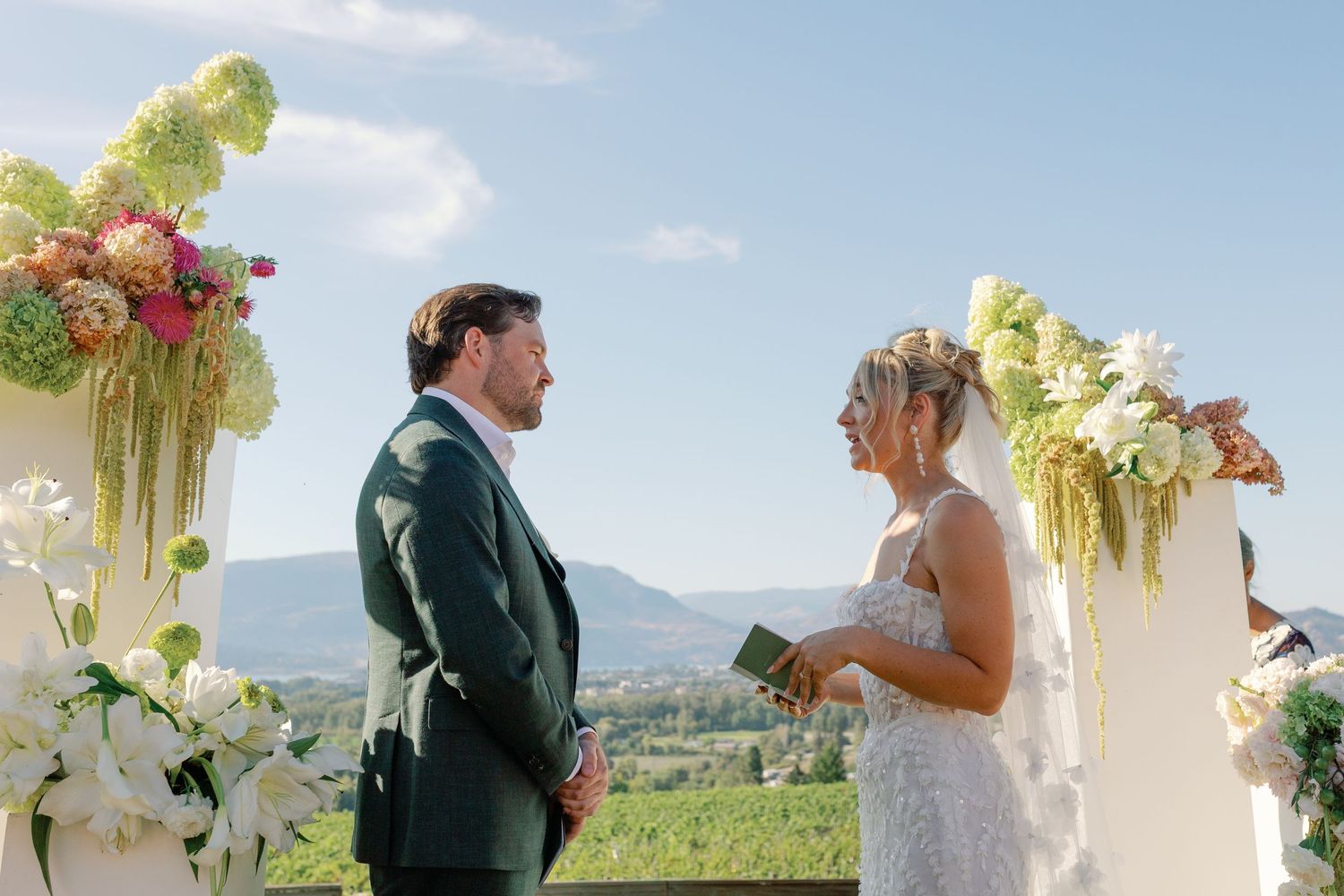 Couple stands at vineyard wedding altar surrounded by lush green and pink floral arrangements.