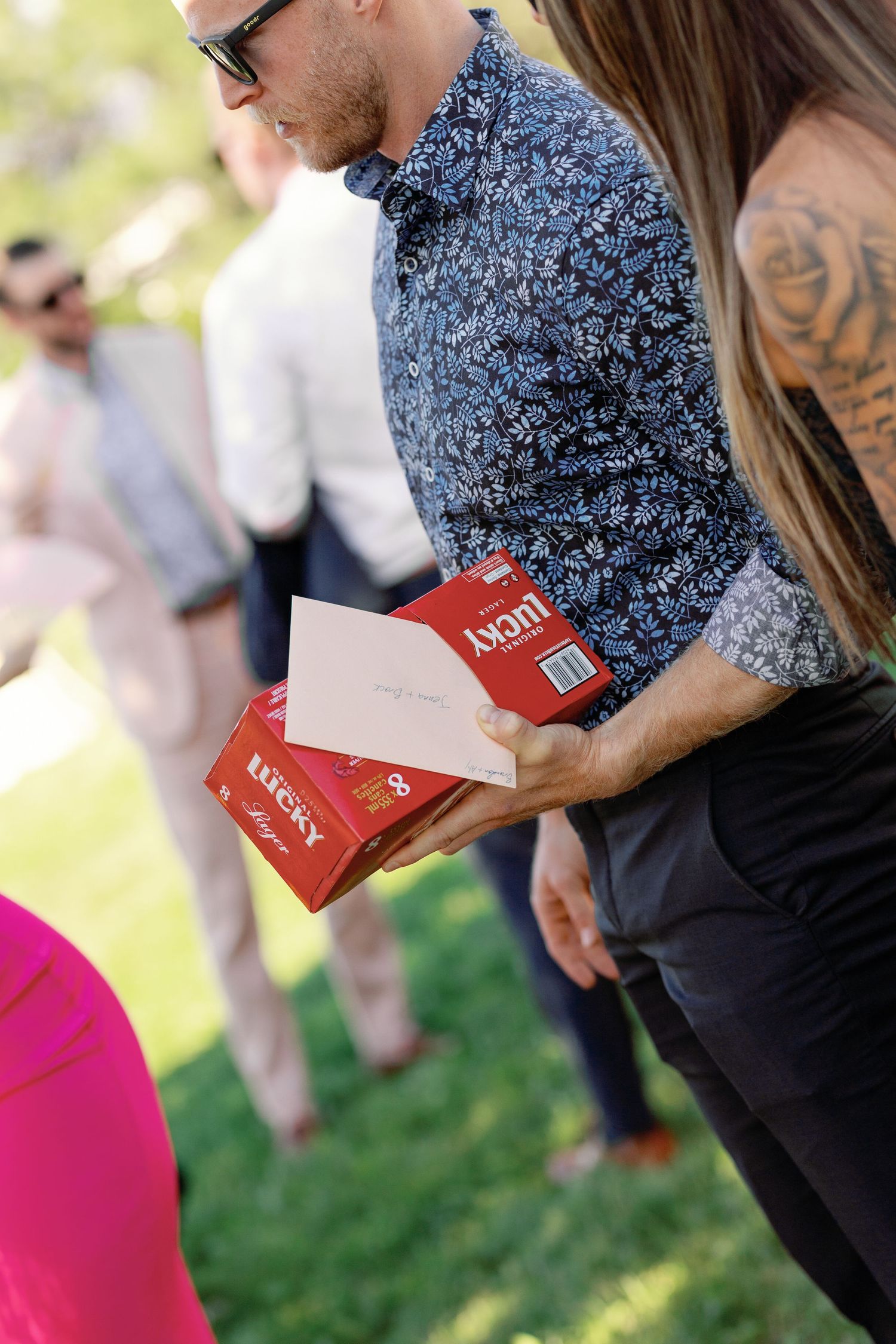 Wedding guest holding Lucky Strike cigarettes and a greeting card at an outdoor ceremony.