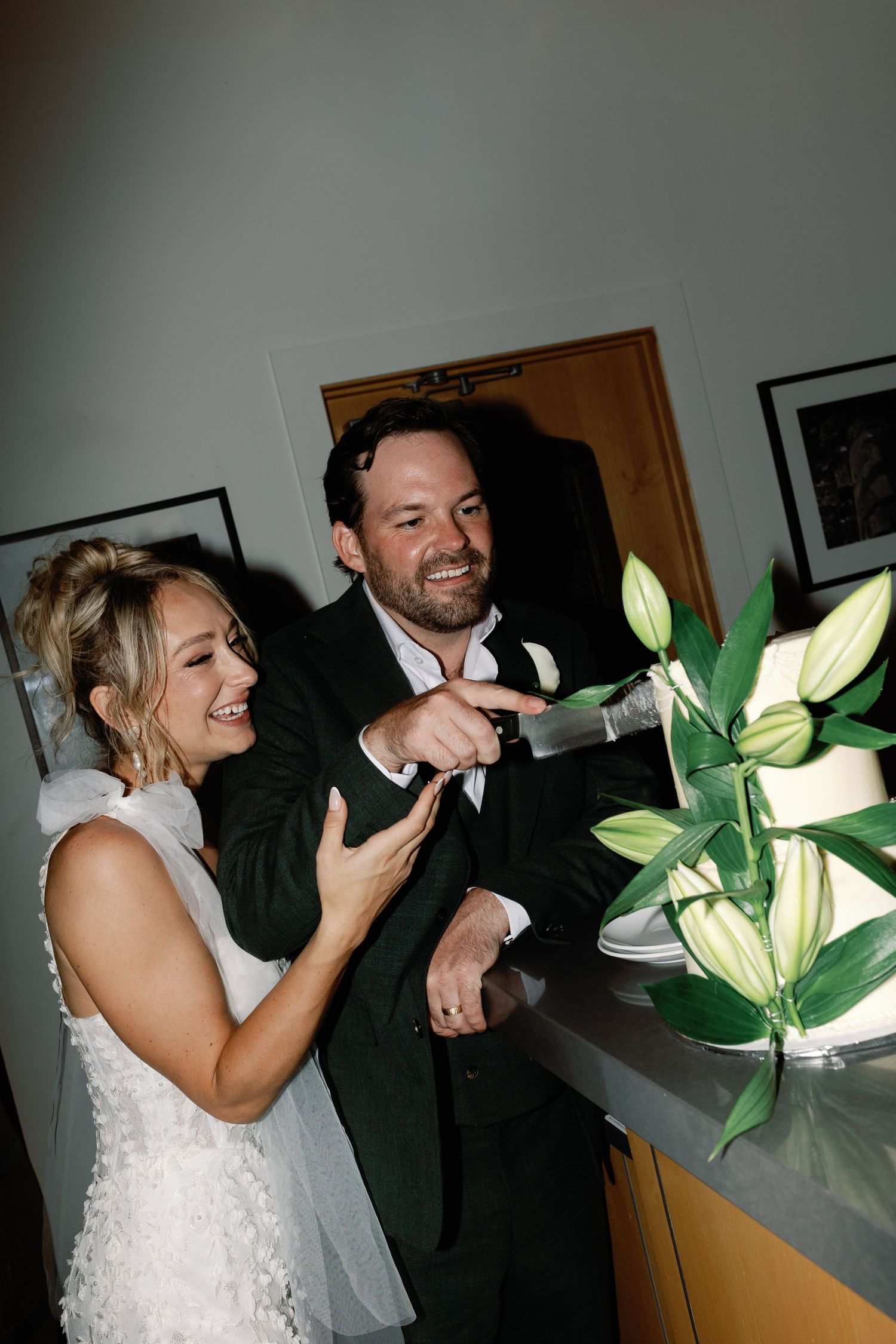 Laughing bride and groom cut their white lily-decorated wedding cake together indoors.