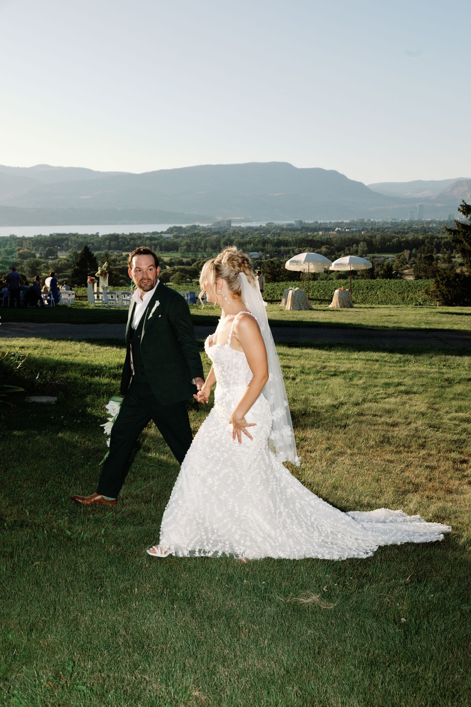 Bride and groom walking on lush green lawn with stunning mountain and lake backdrop at sunset.