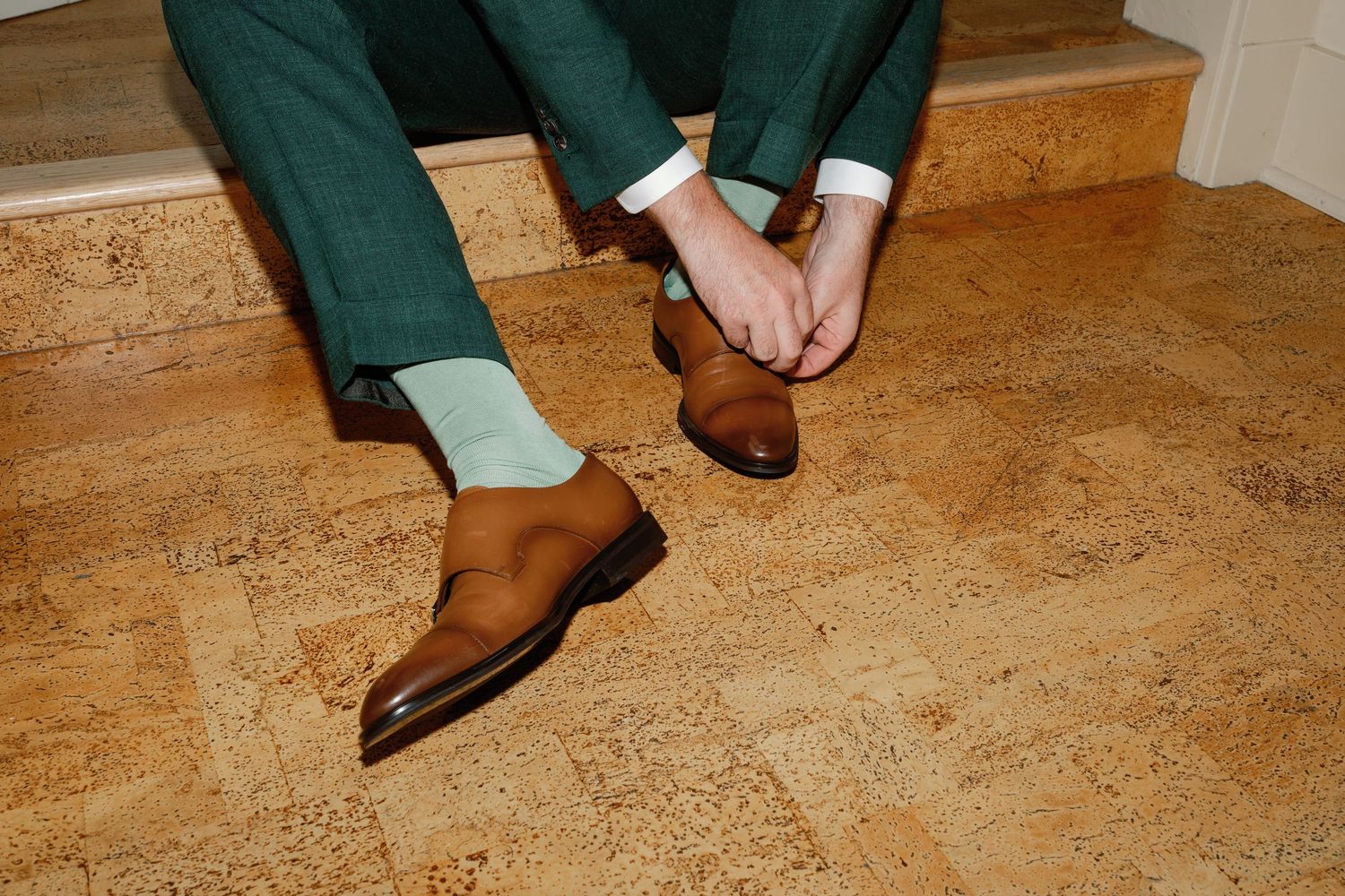 Groom in green trousers and mint socks fastens brown oxford shoes while seated on cork floor steps.