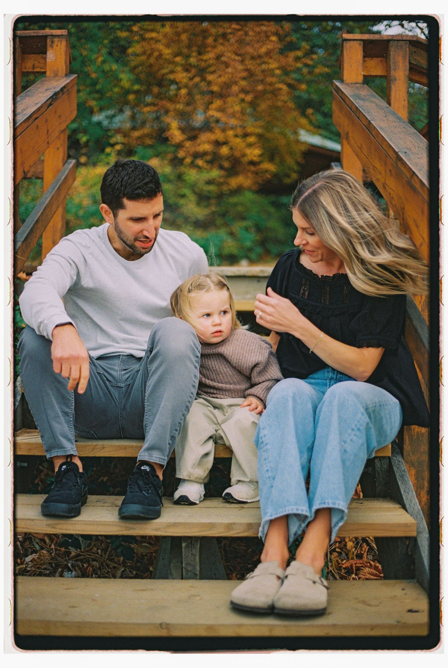 A young family sits on wooden bridge steps surrounded by autumn foliage, with a toddler standing between his parents.