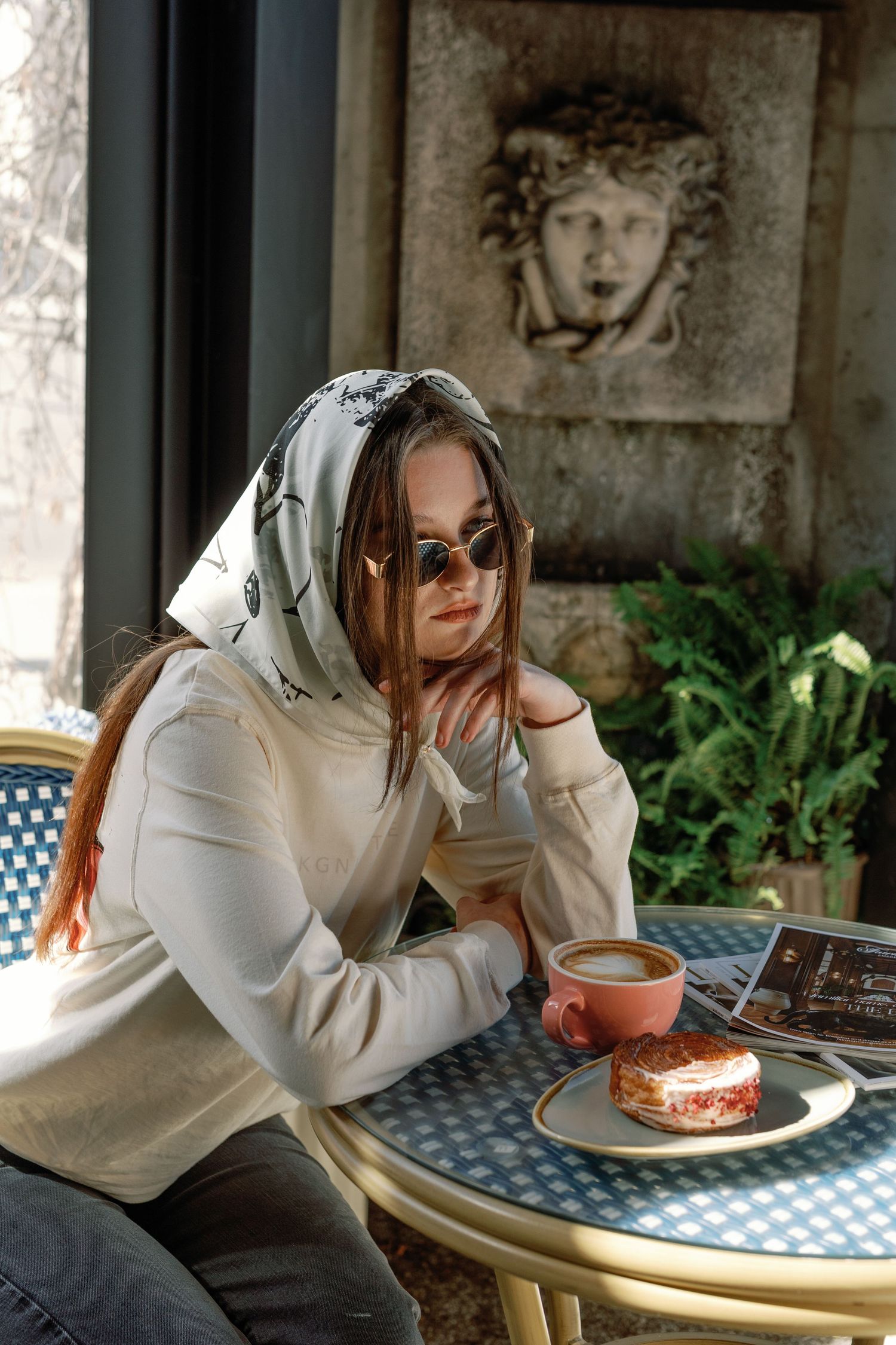 Young woman in white hoodie and headscarf sits at cafe table with coffee and pastry, stone relief on wall behind her.