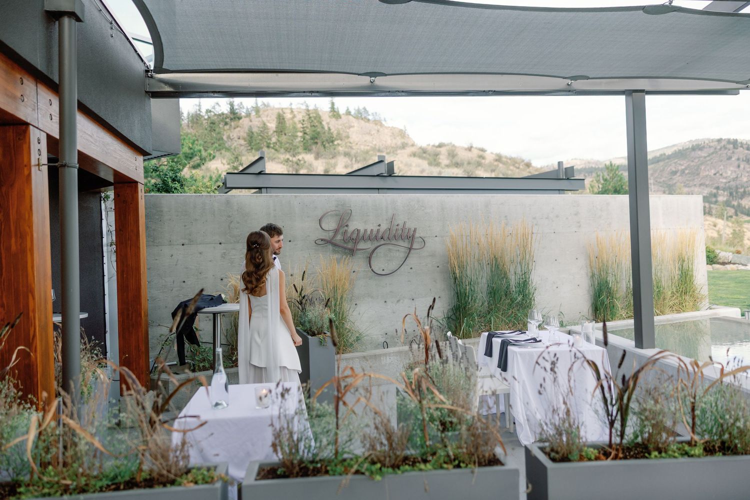 Bride stands on patio of Liquidity Winery restaurant with mountain landscape in background.