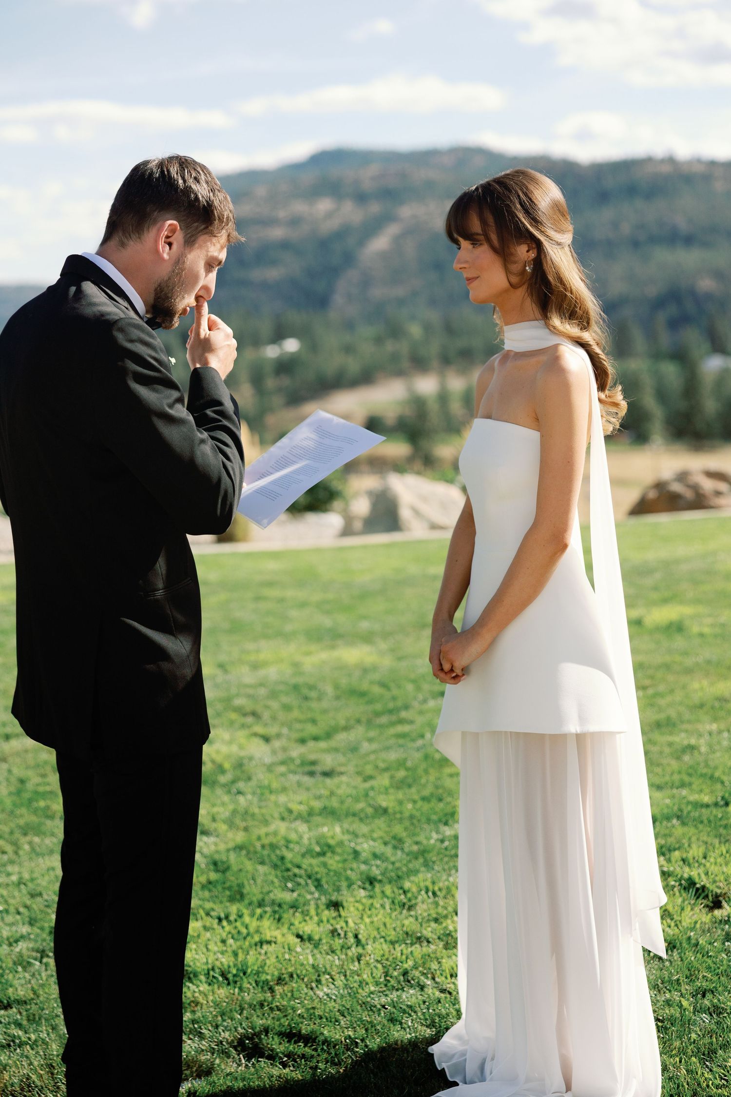 Emotional groom reads handwritten vows to bride during outdoor wedding ceremony with mountain views behind them.
