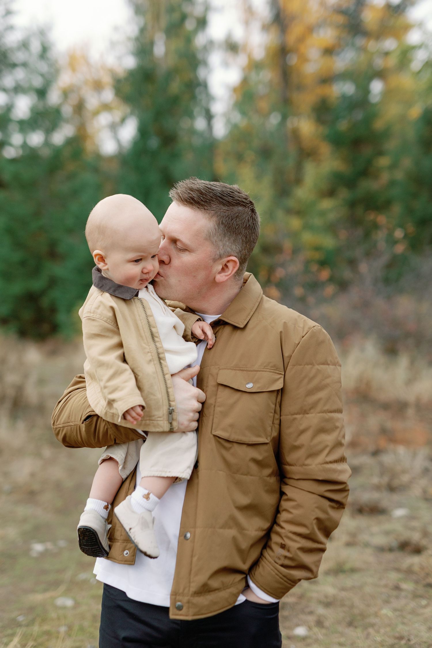 A father in a tan jacket kisses his smiling baby outdoors in an autumn forest setting.