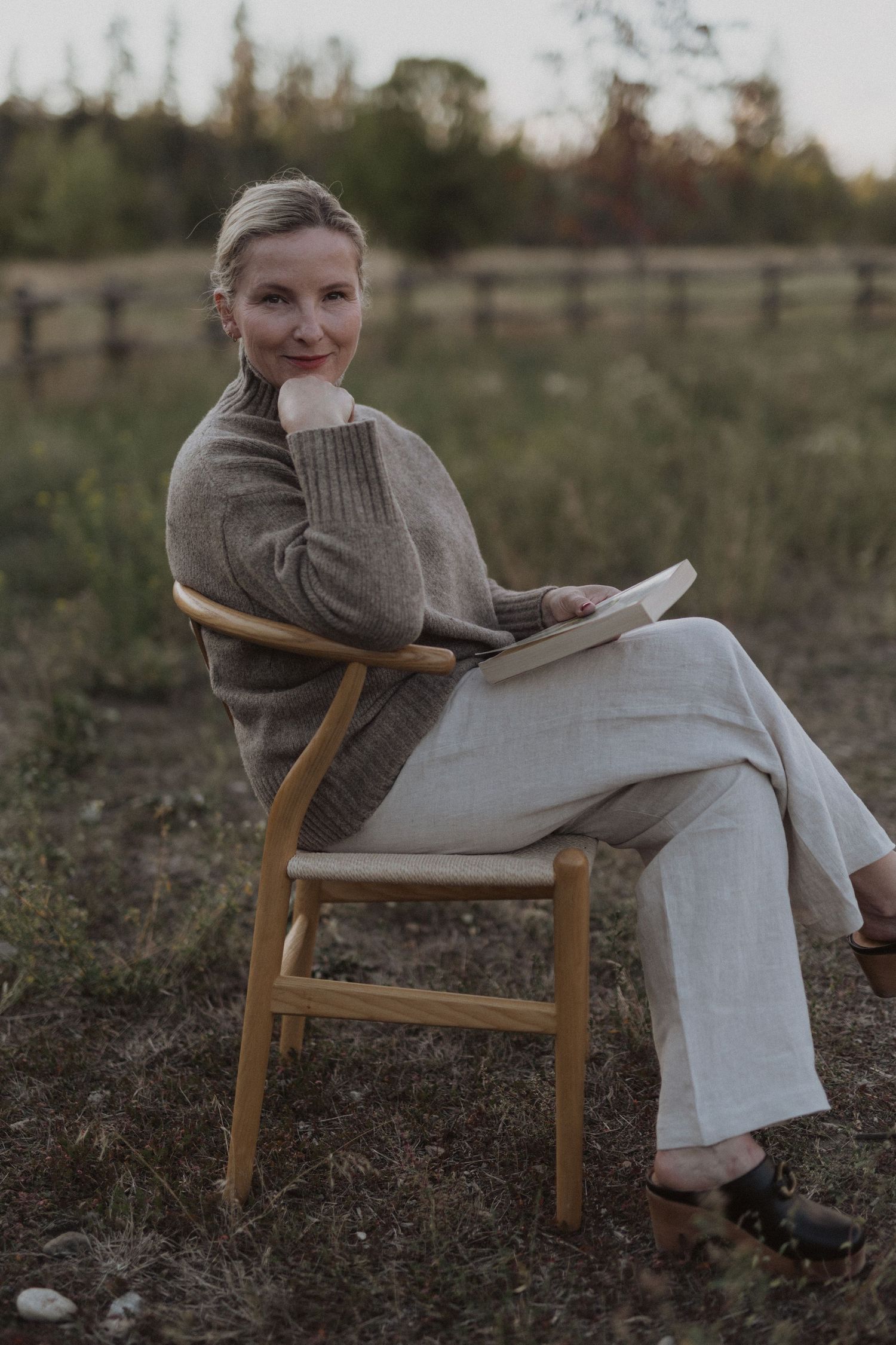 A woman sits relaxed in a wooden chair outdoors, holding a book, wearing a cozy sweater and linen pants.