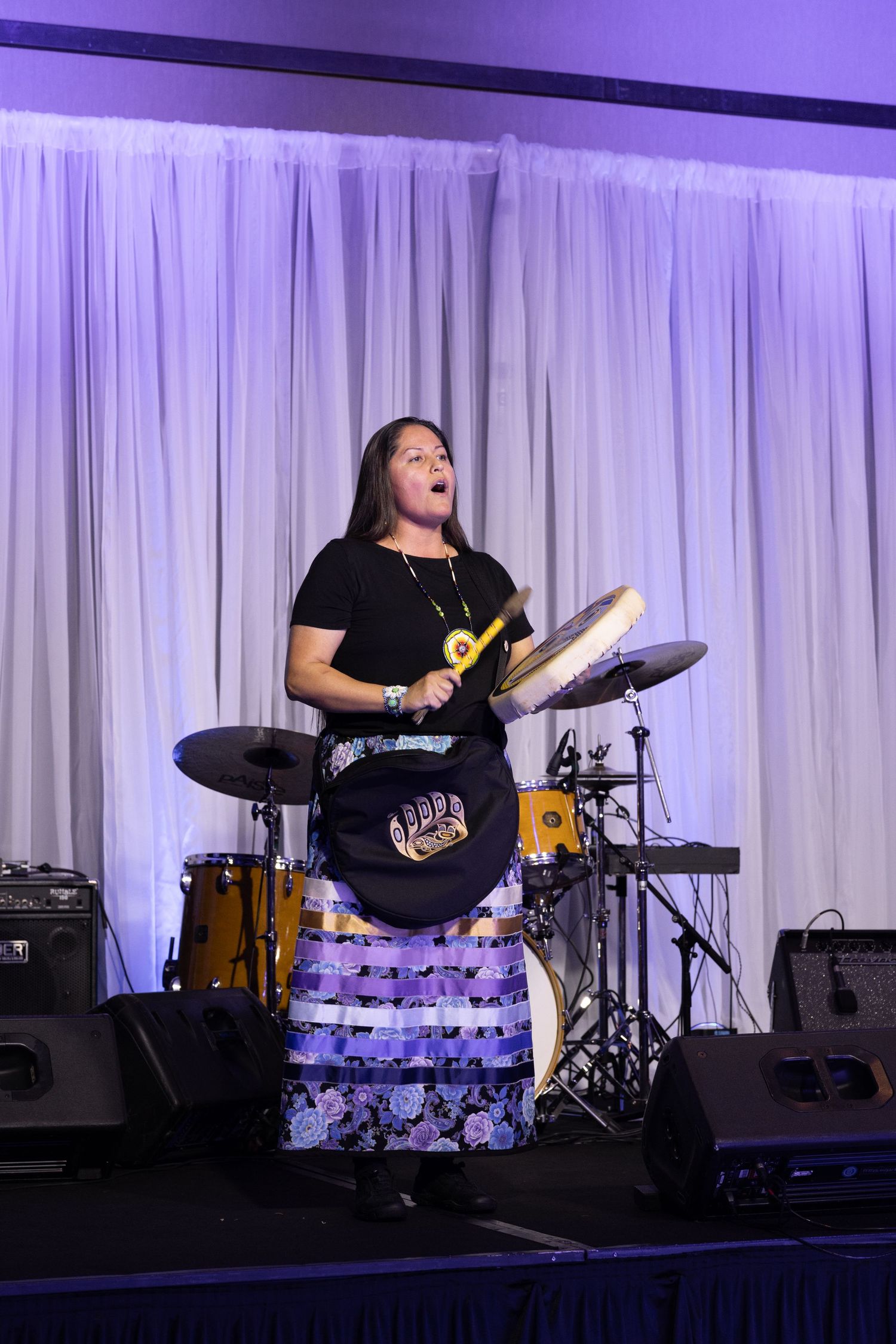 Woman in traditional skirt plays frame drum on stage with purple lighting and white curtain backdrop.