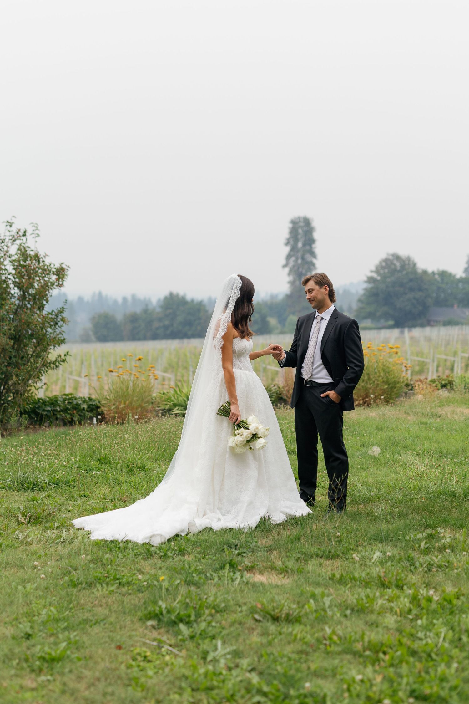 Bride in white gown with veil holds bouquet while groom in dark suit smiles at her in lush green vineyard setting.