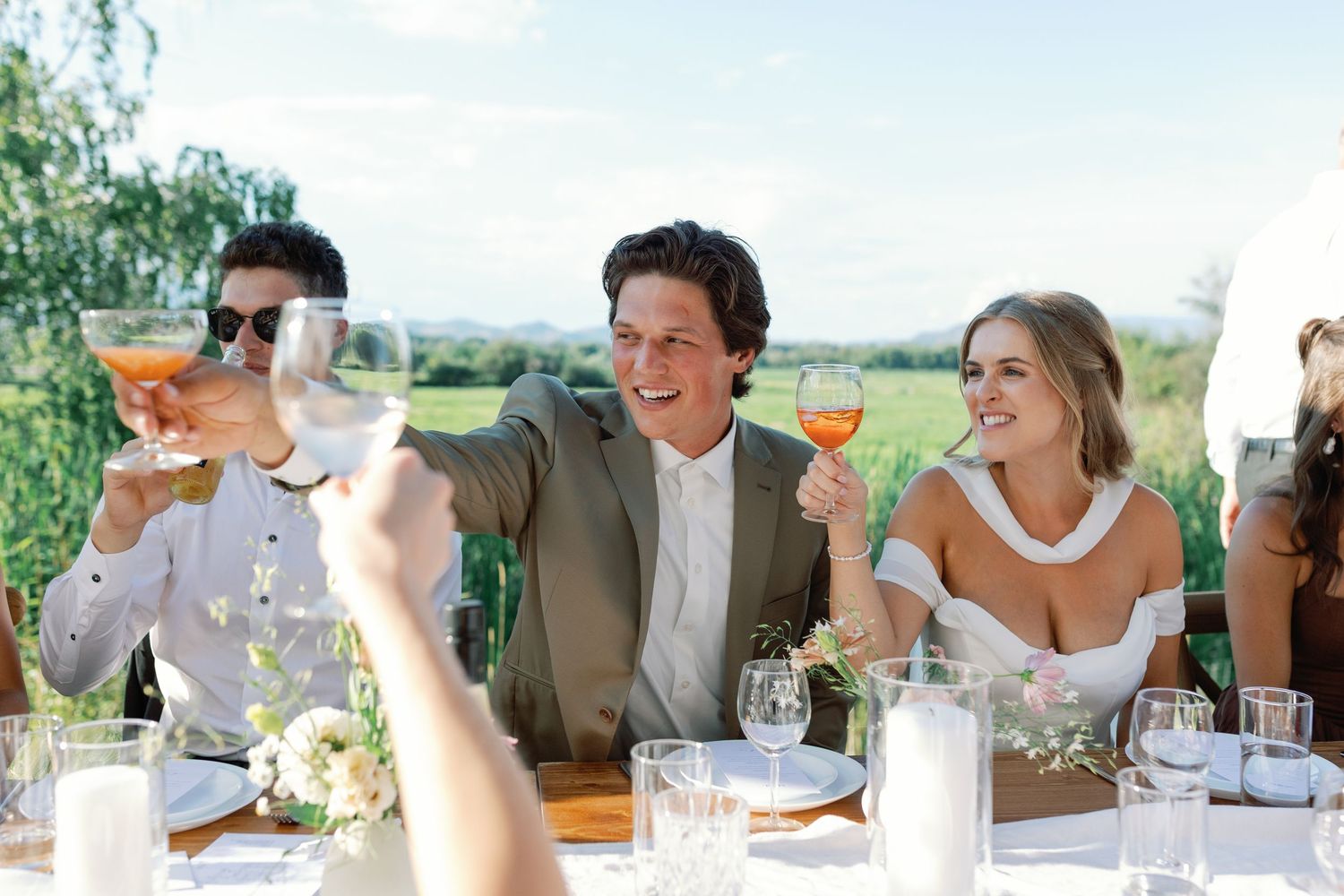 Guests raising glasses in a toast at an outdoor wedding reception surrounded by lush green fields.
