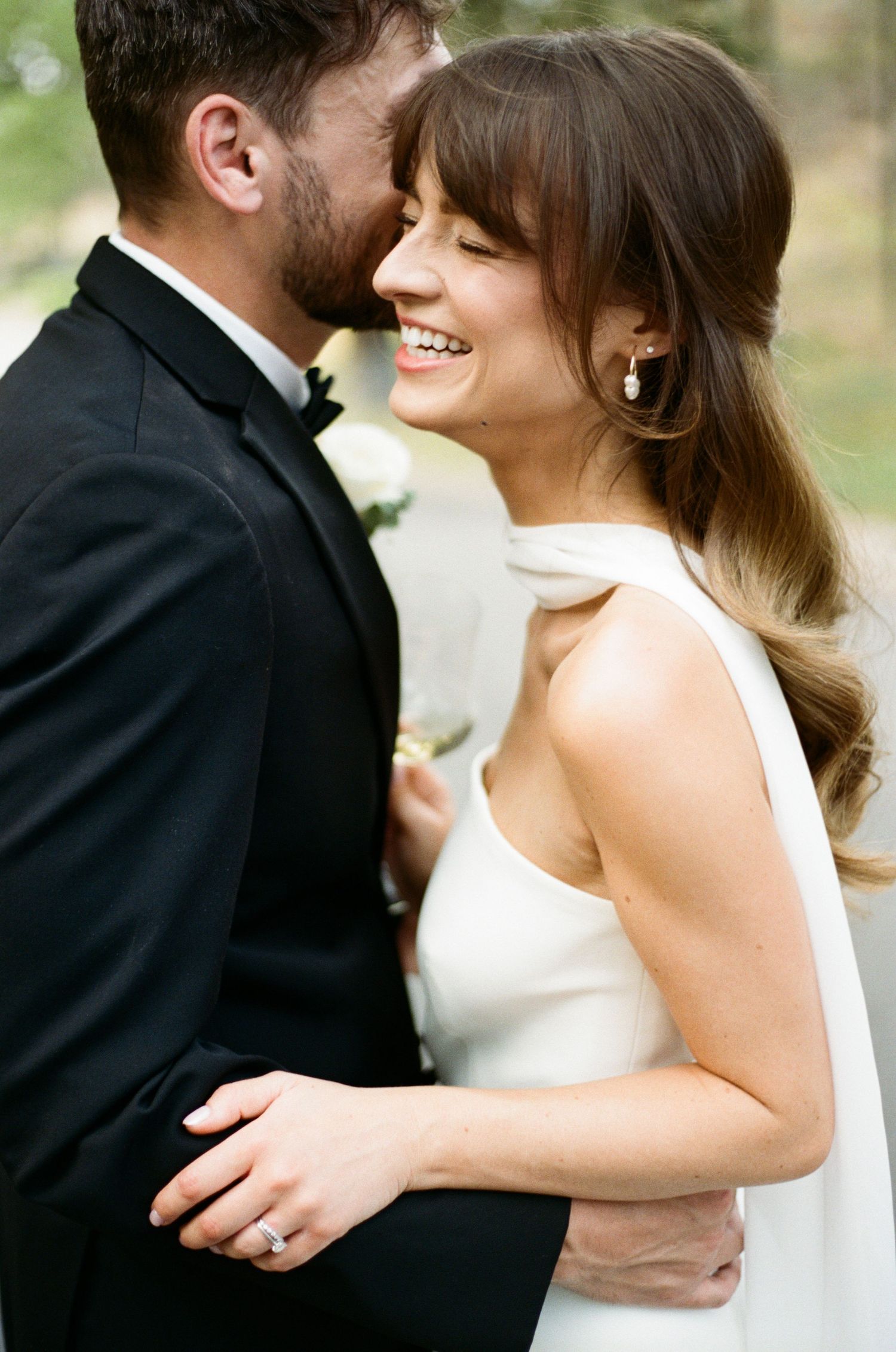 Joyful bride laughing as groom whispers to her, wearing pearl earrings and white halter wedding gown.