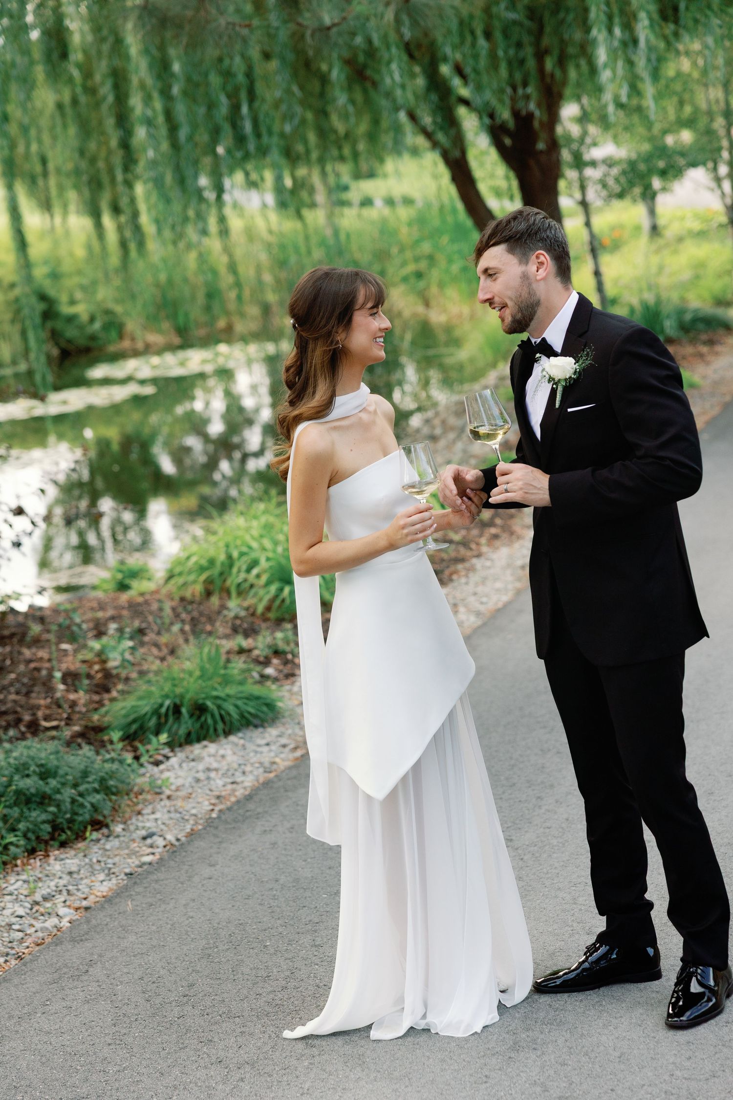 Bride and groom share a romantic moment clinking wine glasses near a serene pond surrounded by weeping willows.